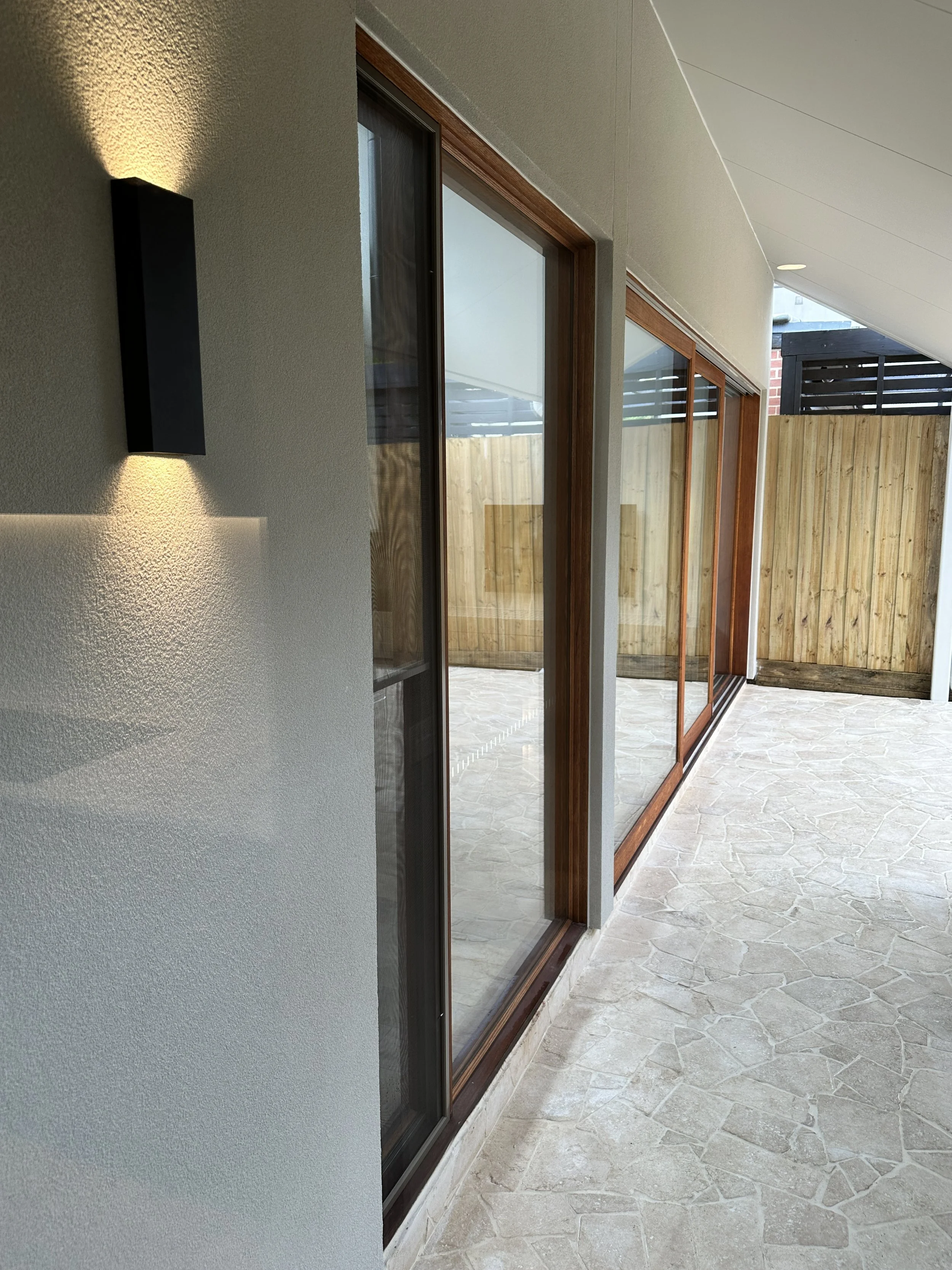 View of a modern house patio with large glass sliding doors, a beige textured wall with a wall light, and a wooden fence in the background.