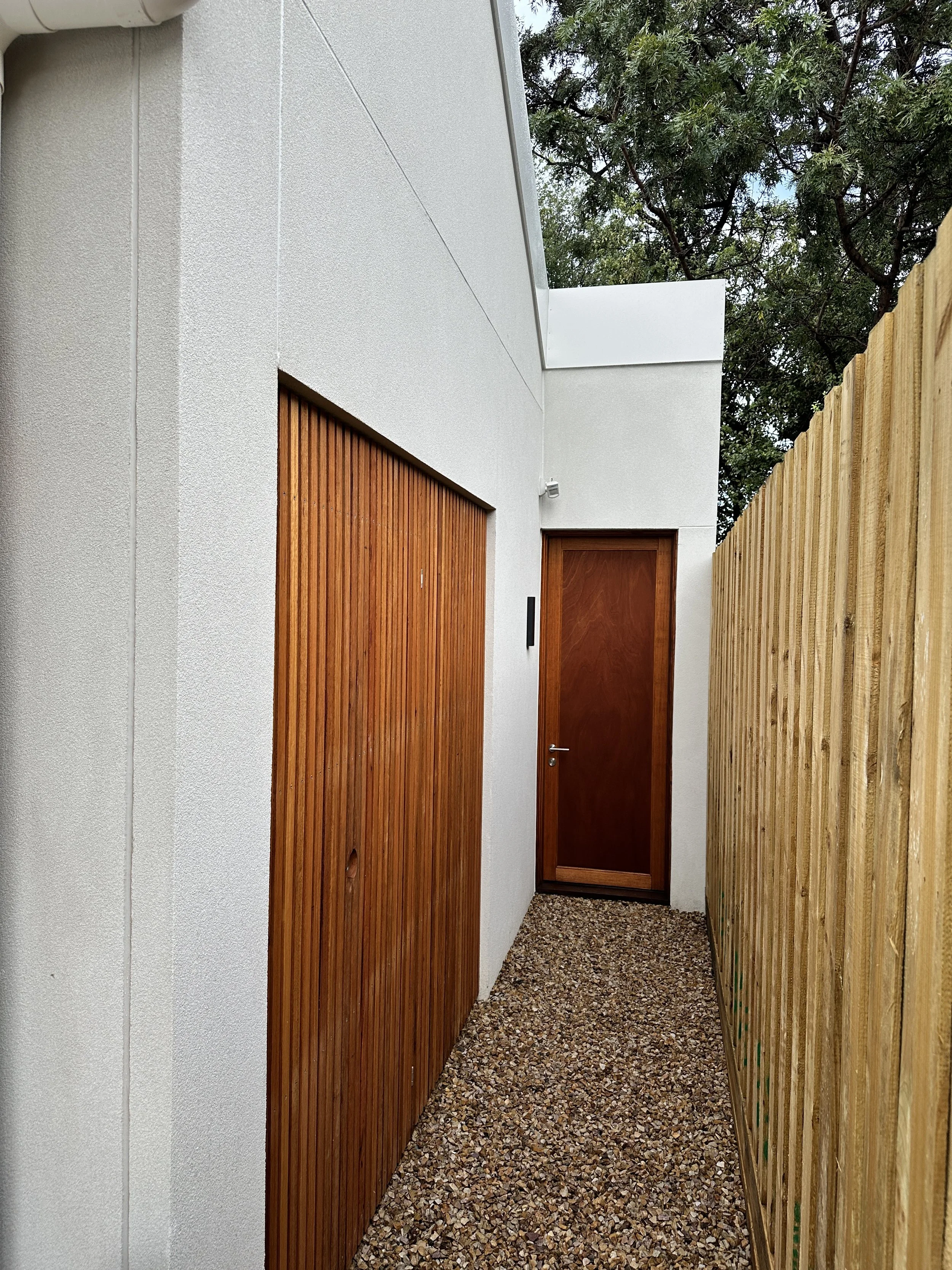 A narrow outdoor passageway with a white building wall on the left, a wooden door and a door with a vertical wood slat design, and a tall wooden fence on the right. The ground is covered with small gravel, and there are trees and a partly cloudy sky 