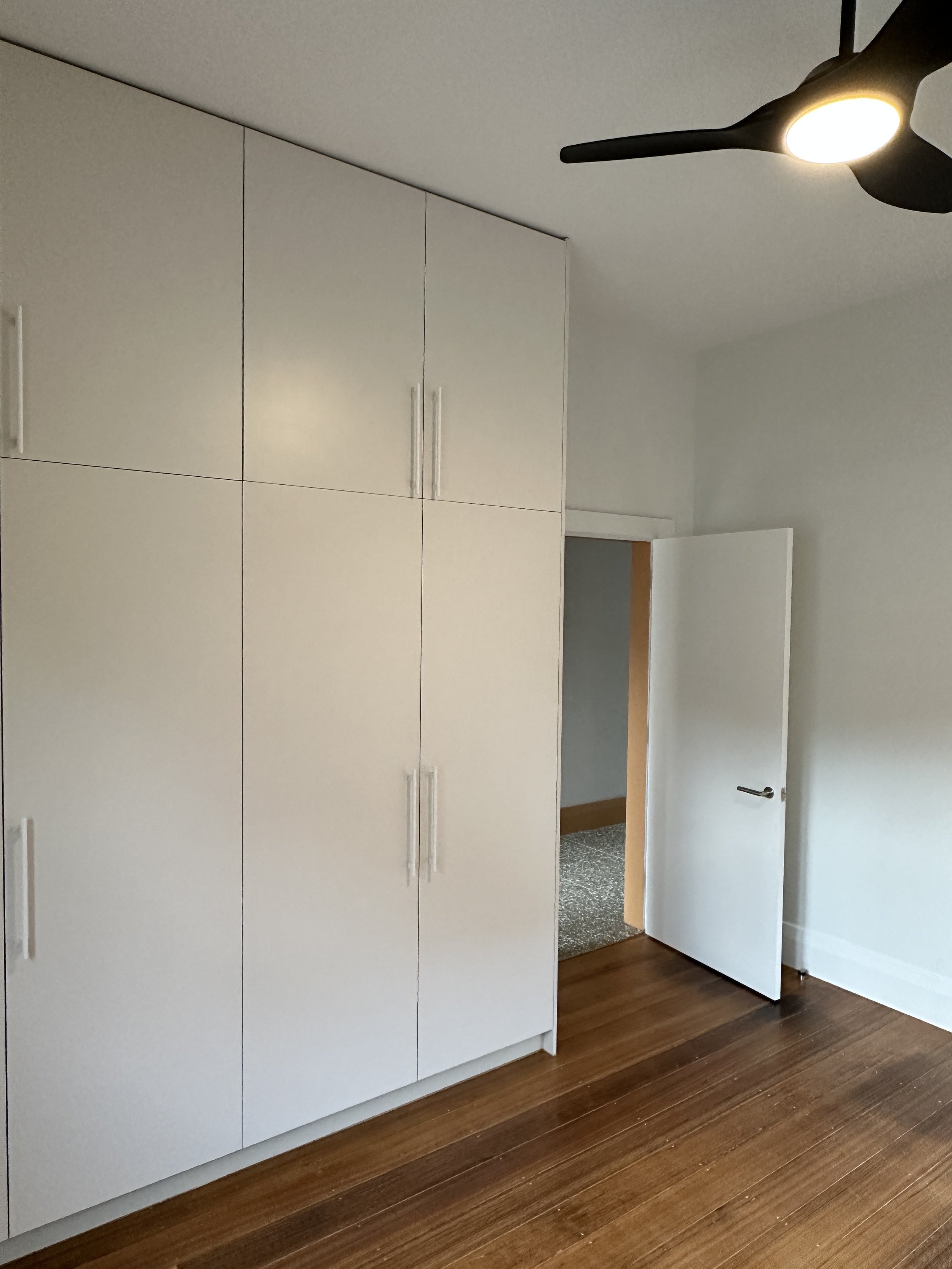 A room with a white wardrobe, a partially open door, hardwood flooring, and a ceiling fan with lights.