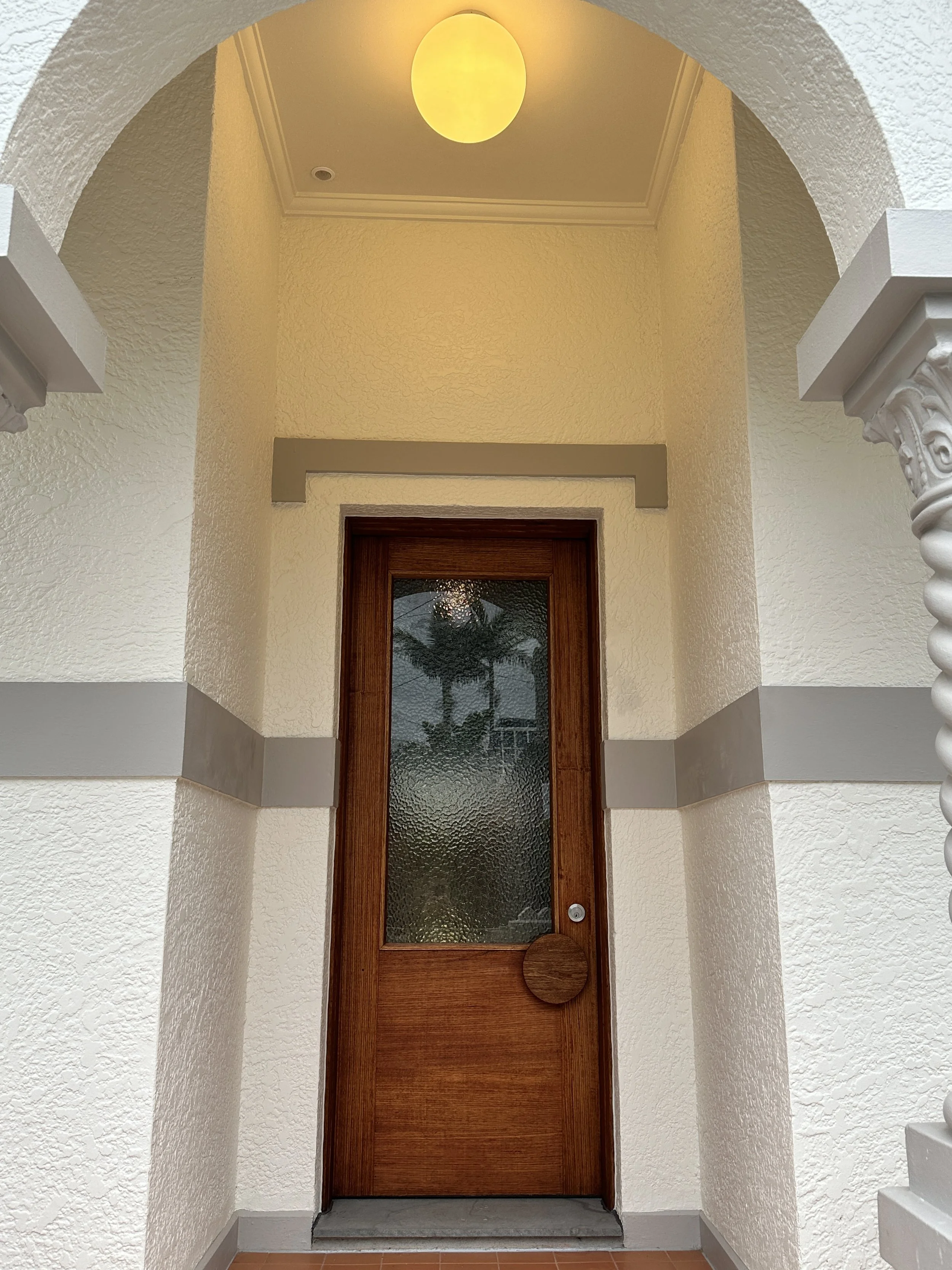 Wooden door with textured glass panel and round wooden handle, set within a white stucco porch with decorative columns and a yellow ceiling light.