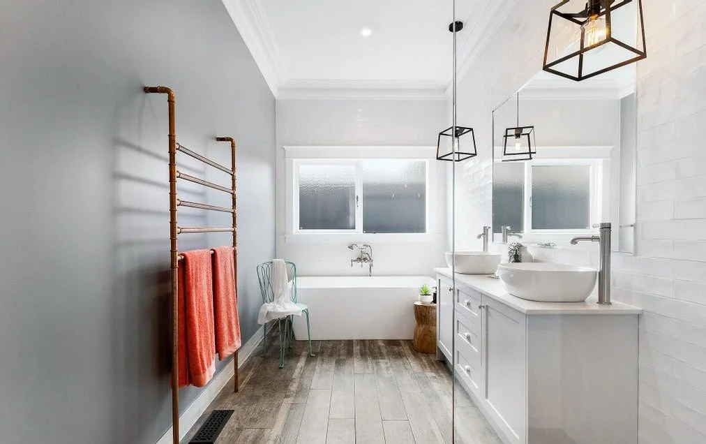 Modern bathroom with white double sink vanity, large window, and bathtub. Brown towel rack with orange towels, decorative pendant lights, and wooden floor.