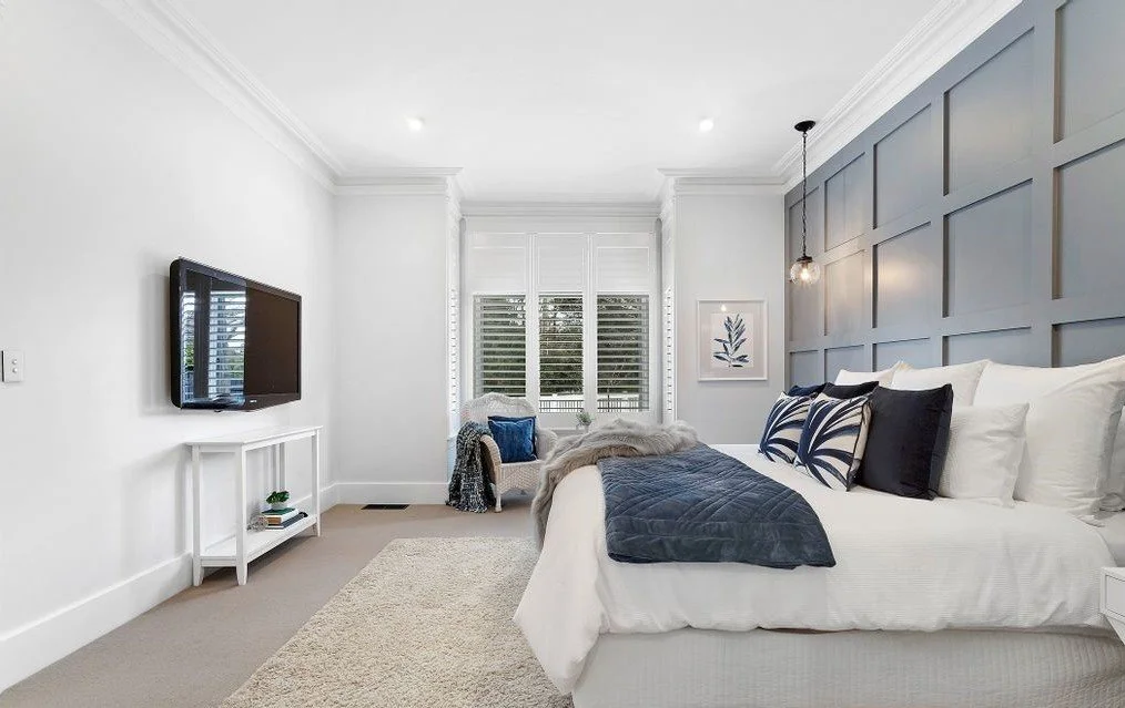 A modern bedroom with white walls and a blue accent wall behind the bed. The room features a large window with white blinds, a white bed with navy and black pillows, a hanging pendant light, a small framed artwork, and a flat-screen TV mounted on the