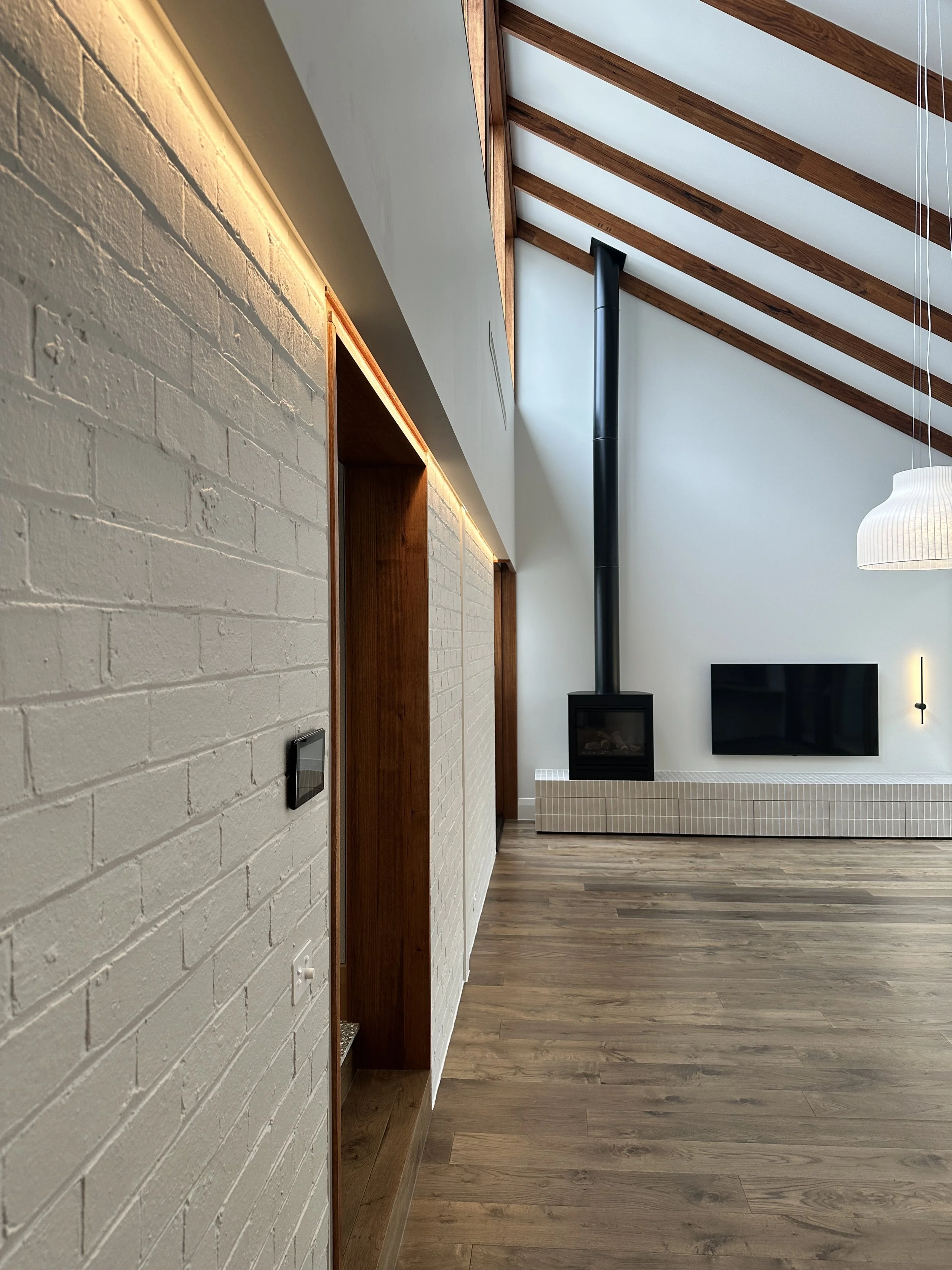 Interior view of a living room with a white brick wall, wooden accents, a built-in electric fireplace with a black chimney, a mounted flat-screen TV, and a sloped wooden ceiling with exposed beams.