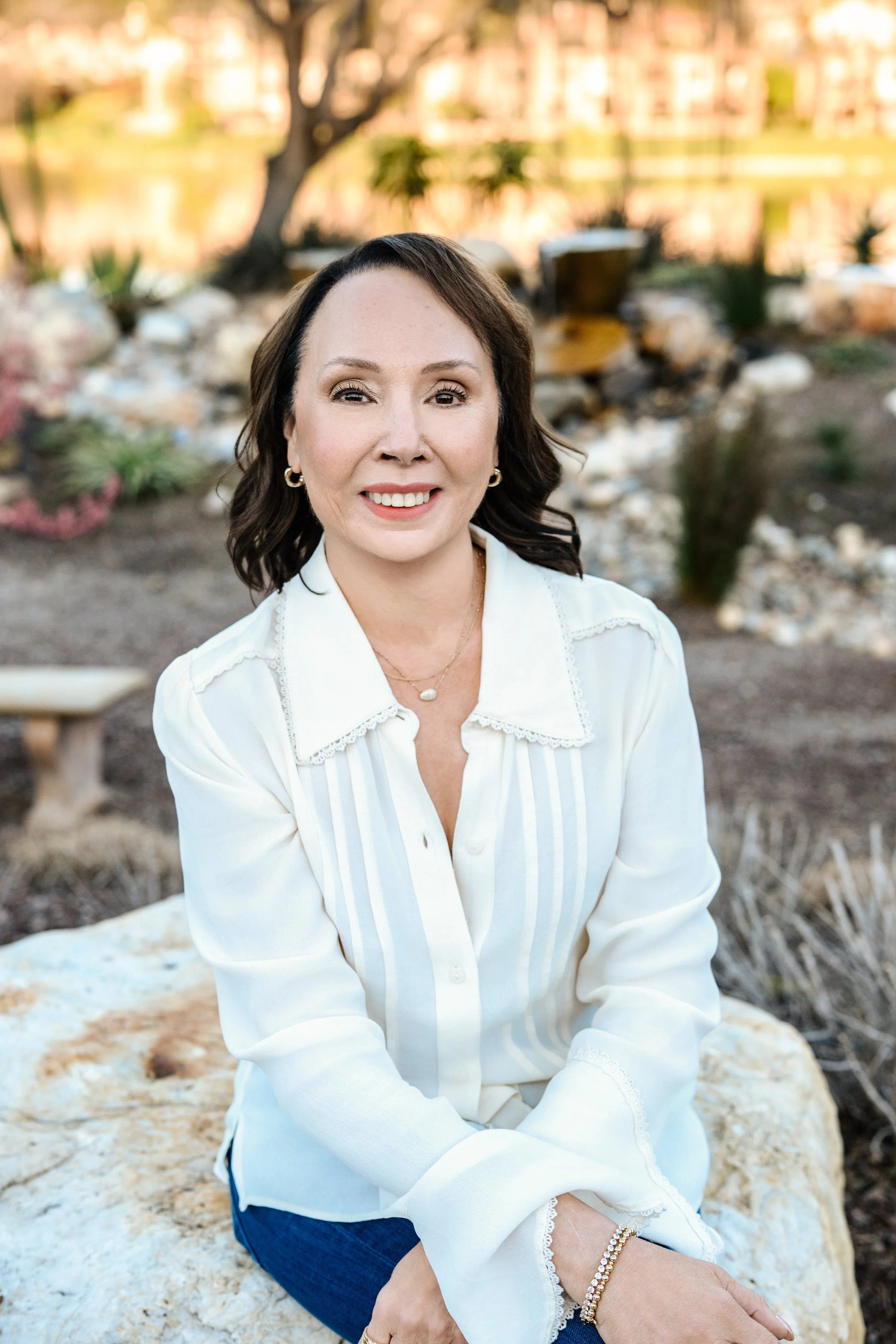 A woman with dark brown hair, wearing a white blouse with lace details, sitting on a large rock outdoors in a garden.