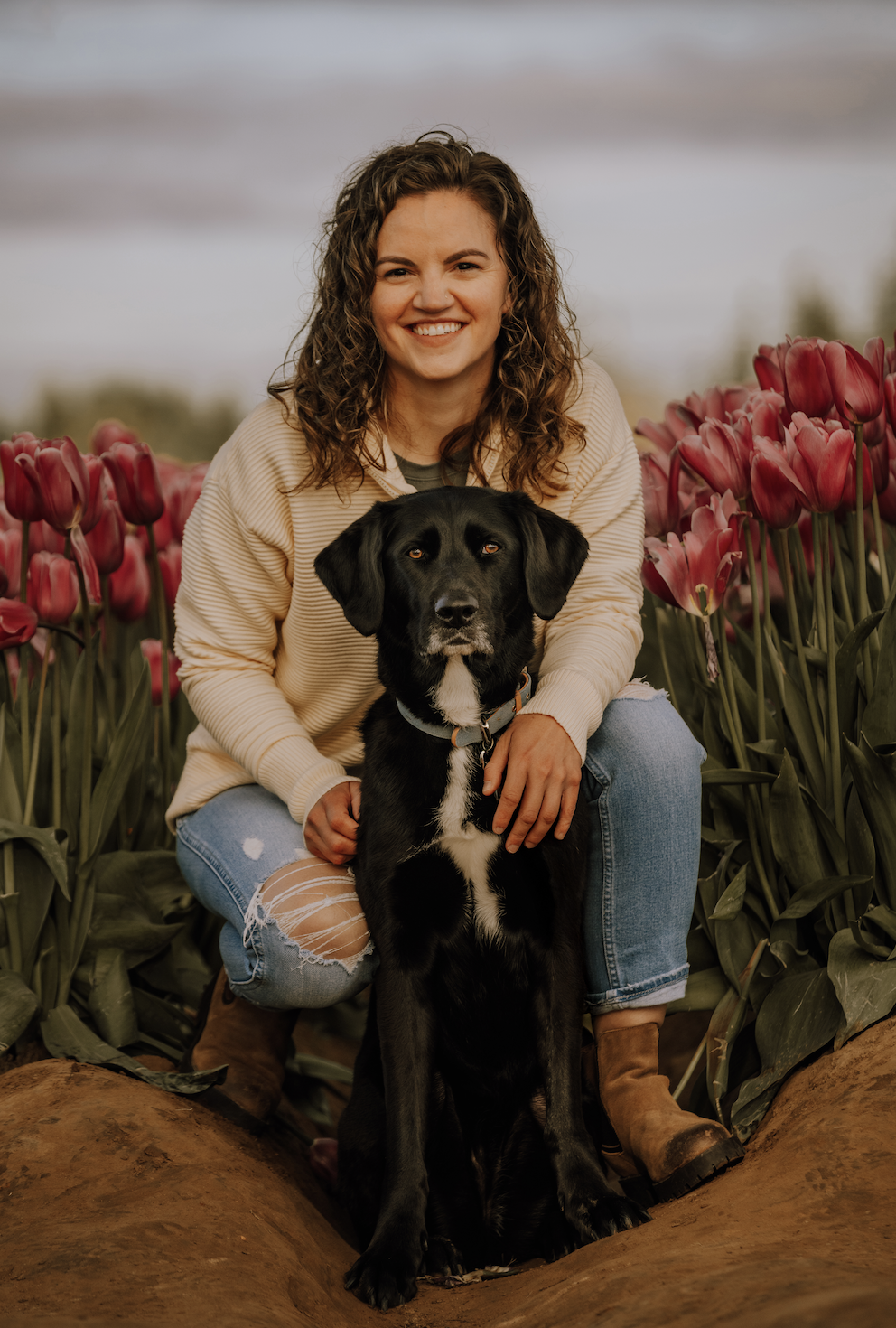 A woman with brown curly hair, wearing a beige sweater and ripped jeans, smiling and crouching in a flower field with pink tulips, holding a black and white dog.