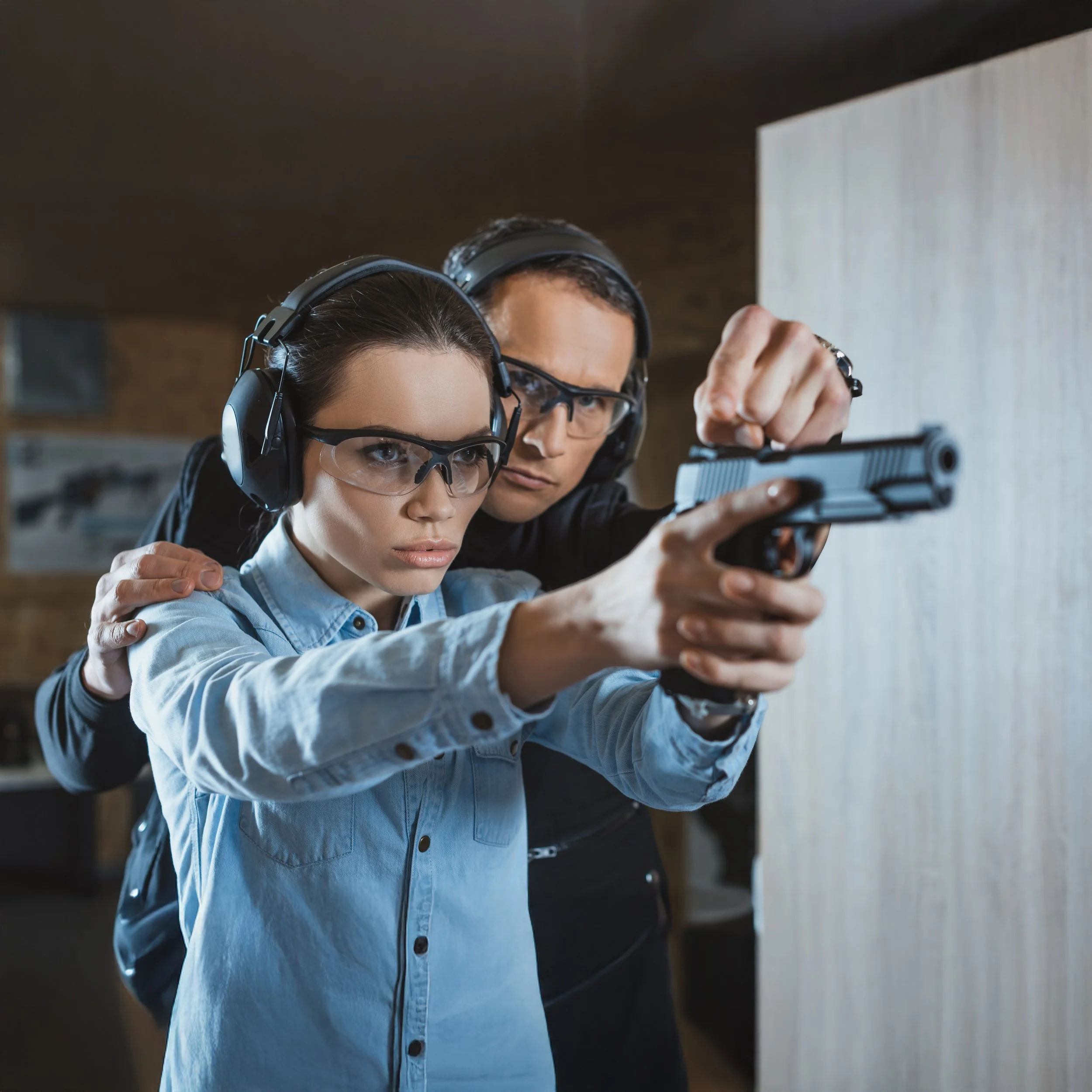 A woman receiving firearm training from an instructor in a shooting range, both wearing safety glasses and ear protection.