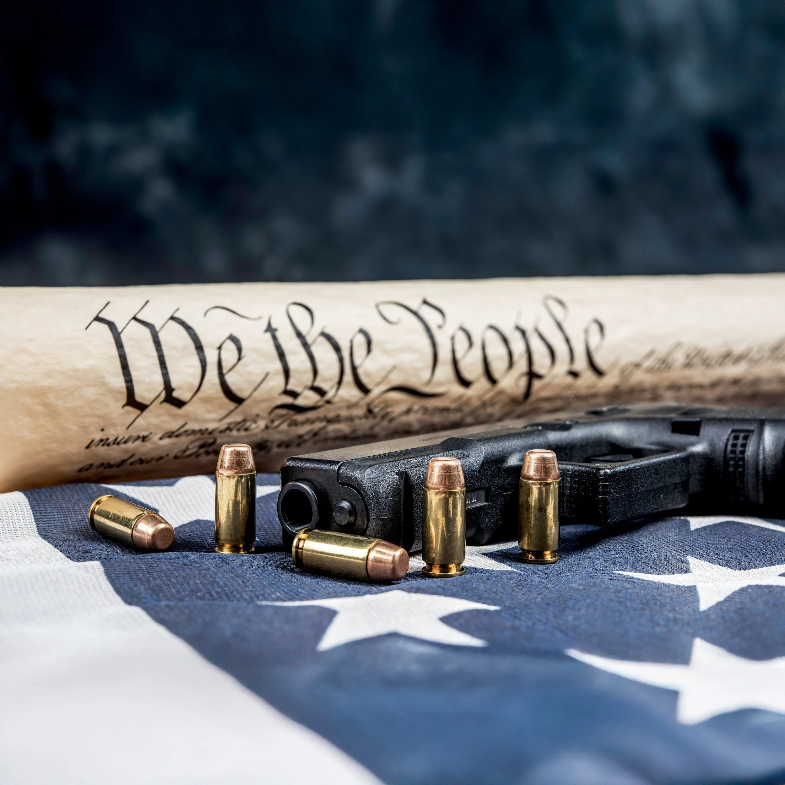 A firearm with five bullets around it, a part of a rolled US Constitution, and an American flag with stars.