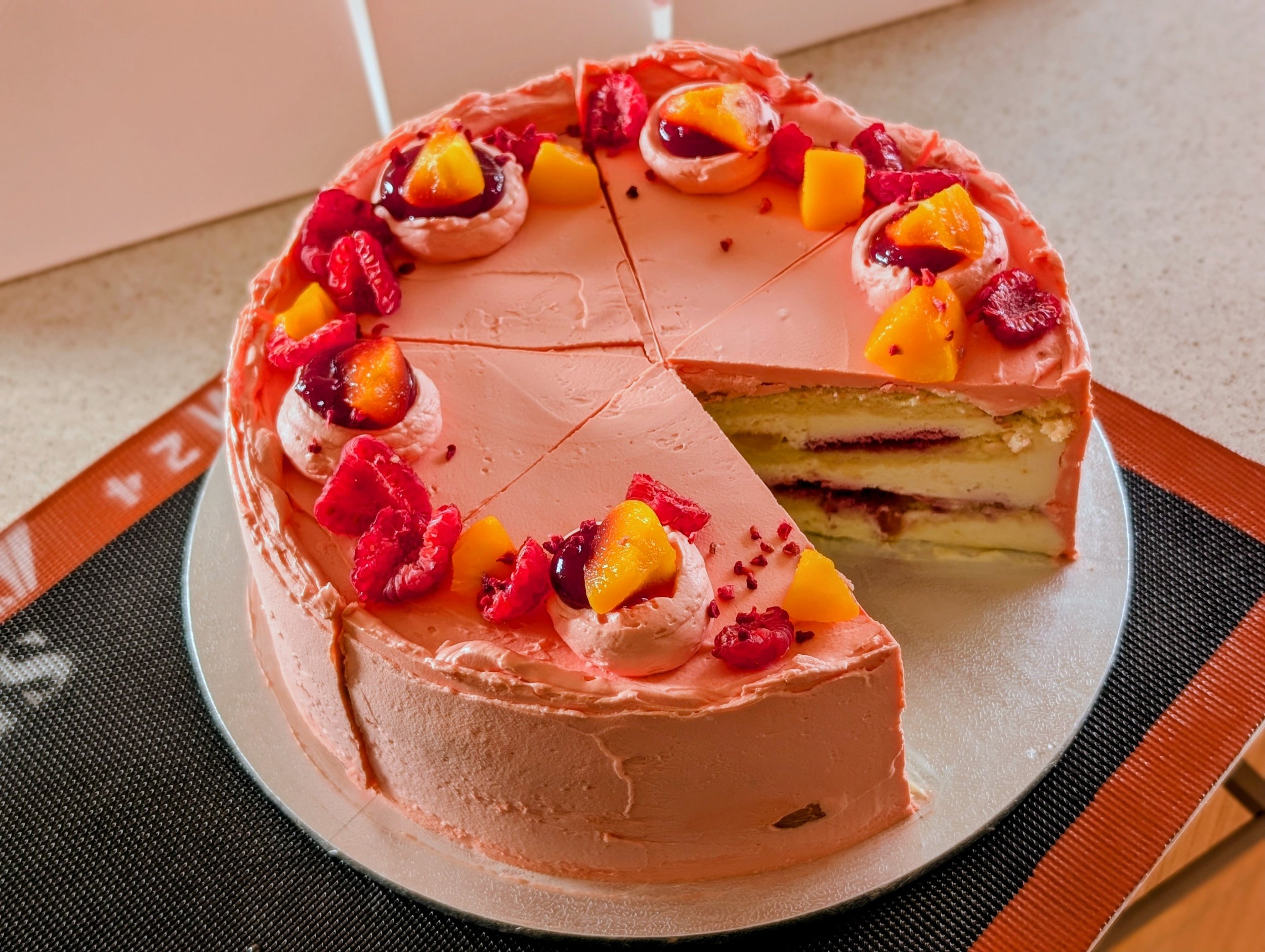 A round layered cake with peach and berry decorations on top, partially sliced to reveal its cream filling and layers inside.