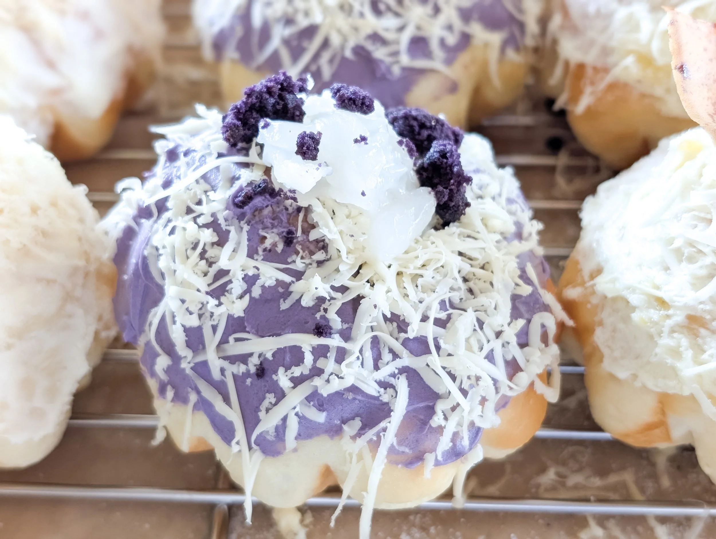 An ensaymada with ube icing topped with white shredded cheese, coconut jelly, and purple crumbles, surrounded by other ensaymadas on a metal rack.