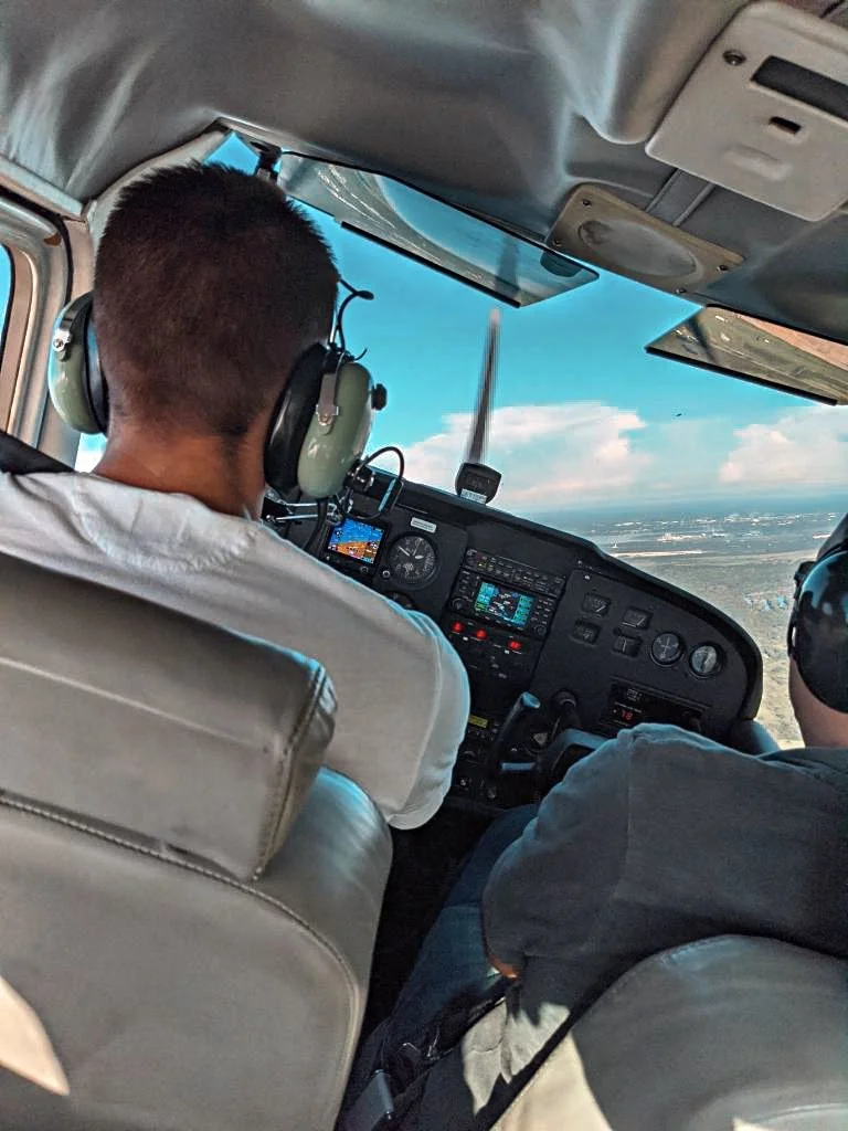 Two pilots flying a small aircraft, viewed from the back seat, with cockpit instruments and a clear sky outside.