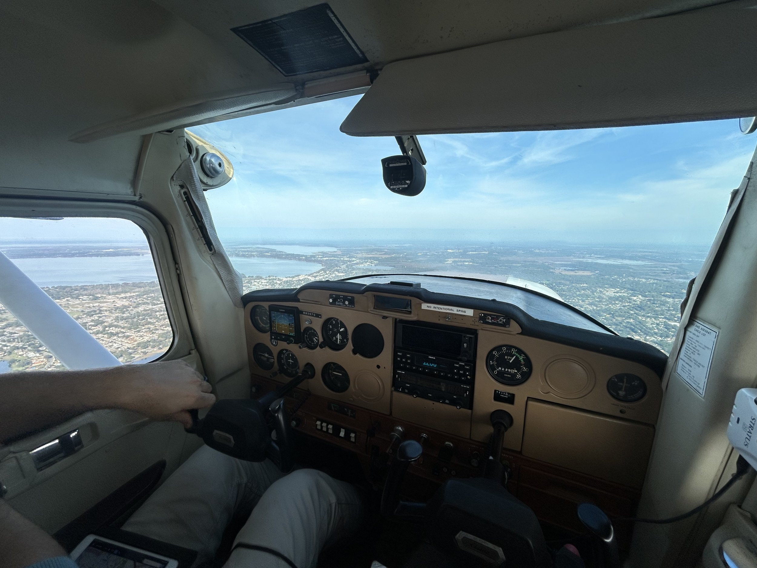 Cockpit of small airplane with pilots' hands on controls, flying over a landscape with water and land visible through the windshield.