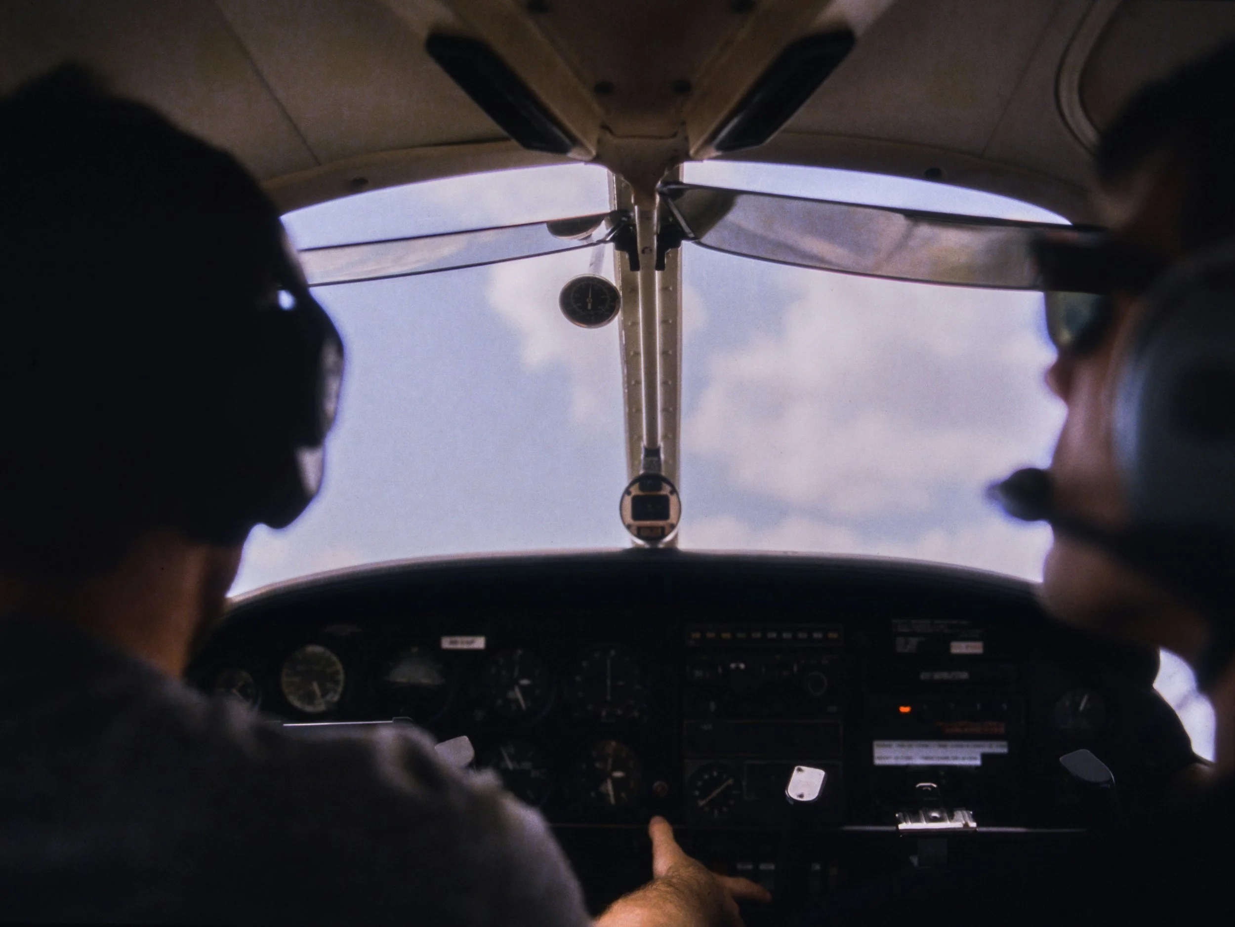Pilot and co-pilot sitting in the cockpit of a plane, viewed from behind, with the sky visible through the windshield.
