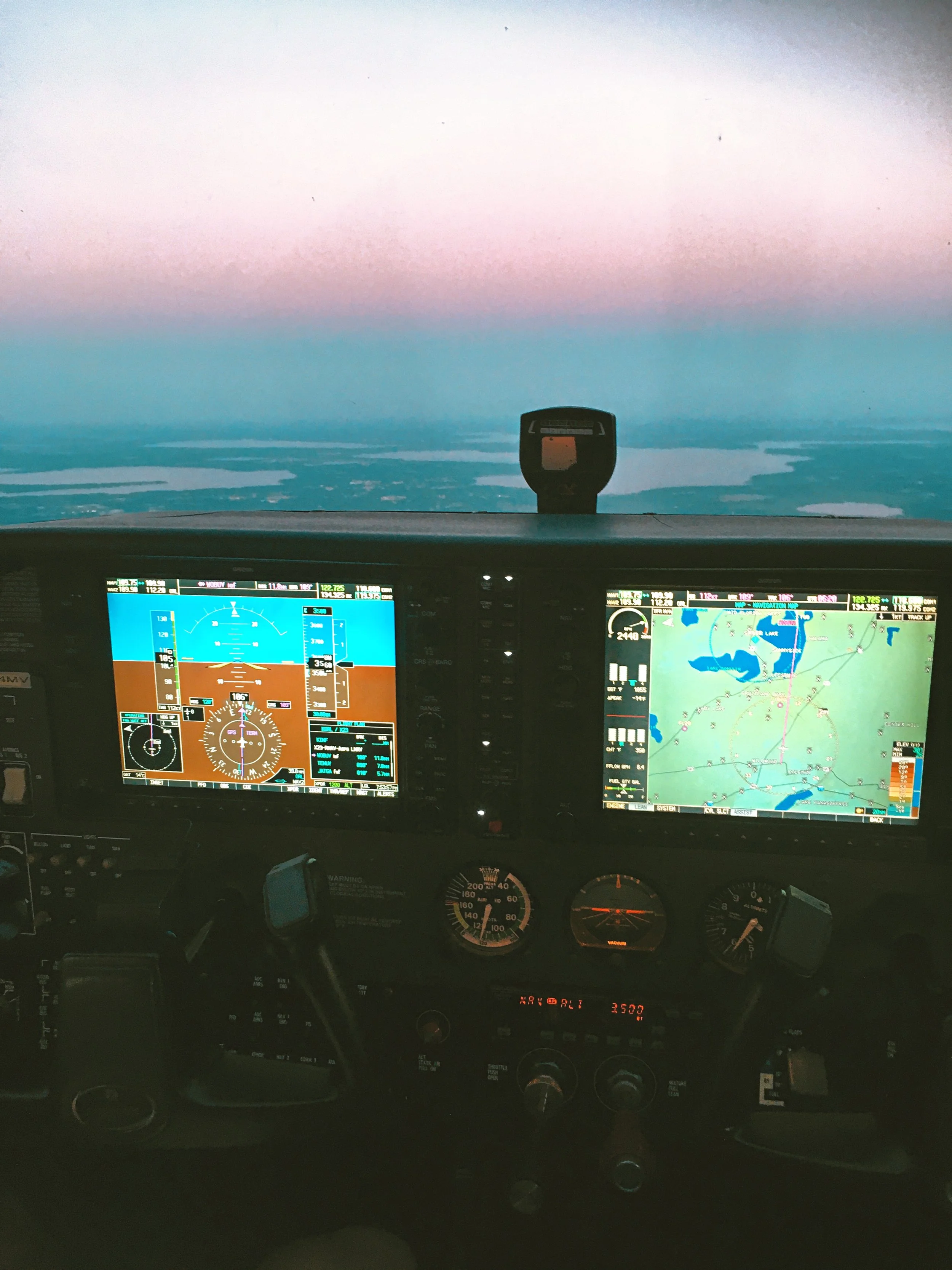 The image shows the cockpit of an airplane with navigation screens, gauges, and controls, flying over a landscape with lakes and rivers at dusk or dawn.