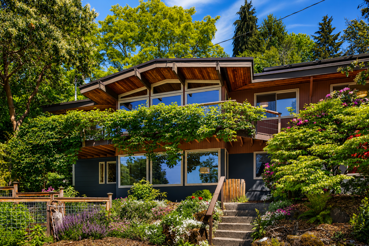 An updated 1962 PNW Seattle modern two-story house with large windows, a balcony, and wooden details, surrounded by lush trees and colorful flowers under a bright blue sky.