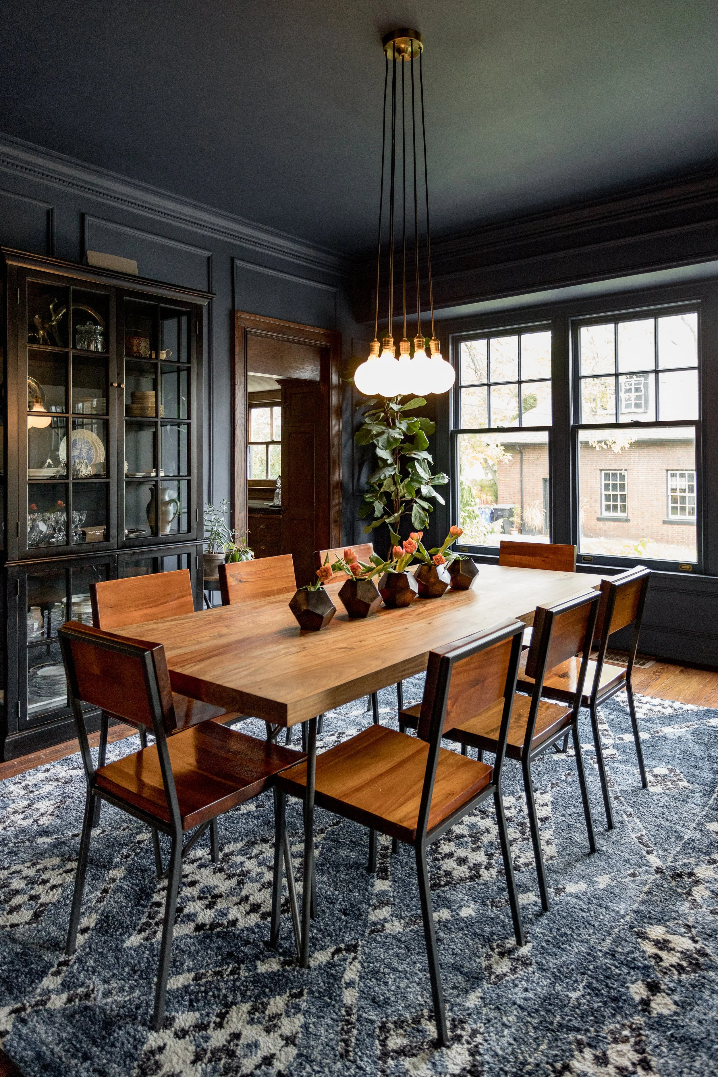 A dining room with a wooden table, surrounded by wooden chairs, black sideboard, and a large nature-inspired rug. A cluster of five globe pendant lights hangs above the table, and a tall houseplant decorates the corner next to large windows that reveal a brick building outside.