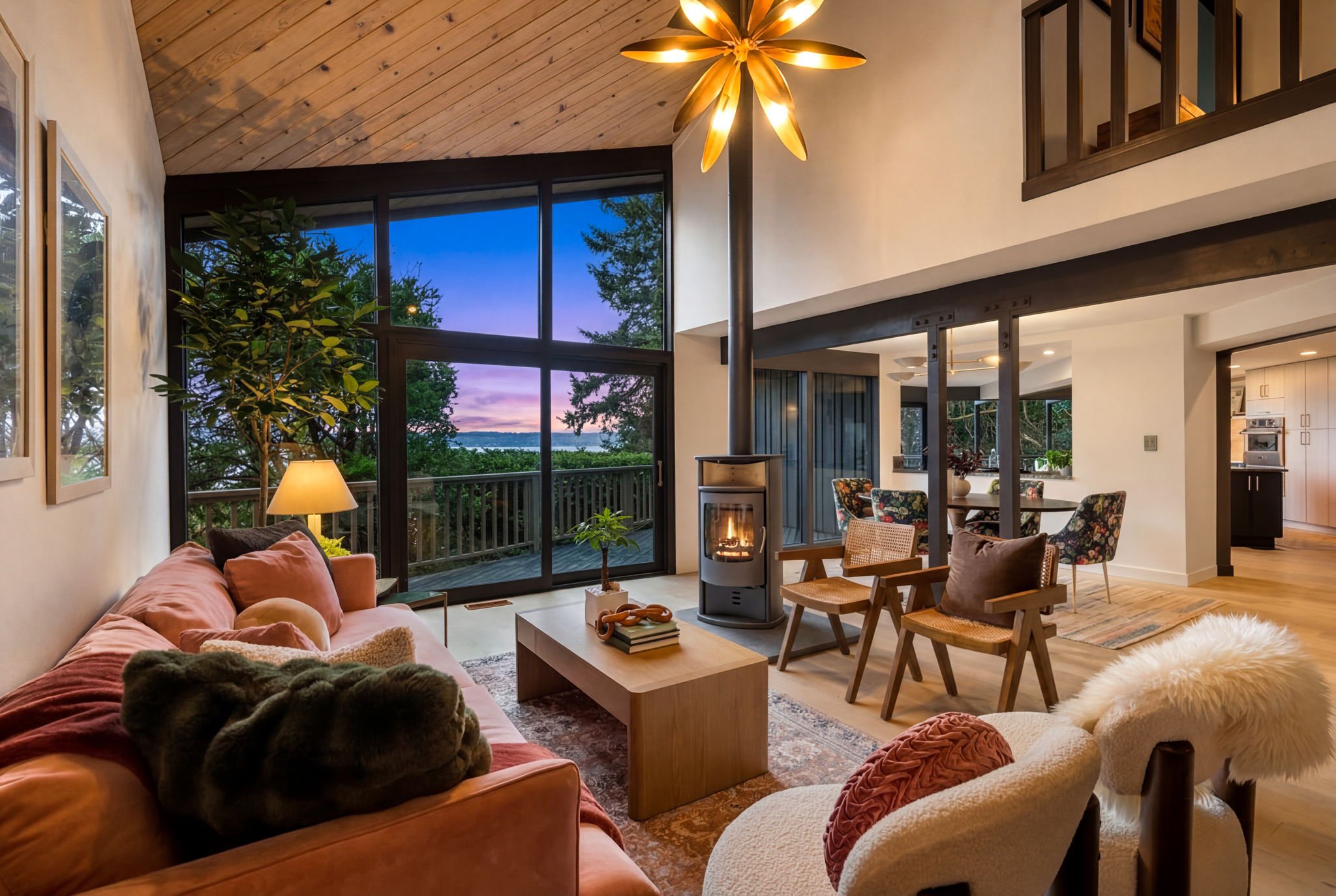 1969 PNW contemporary living room with vaulted wood clad ceiling, architectural chandelier, wood burner, and a wall of windows framing a view of trees and colorful twilight sky over Puget Sound.