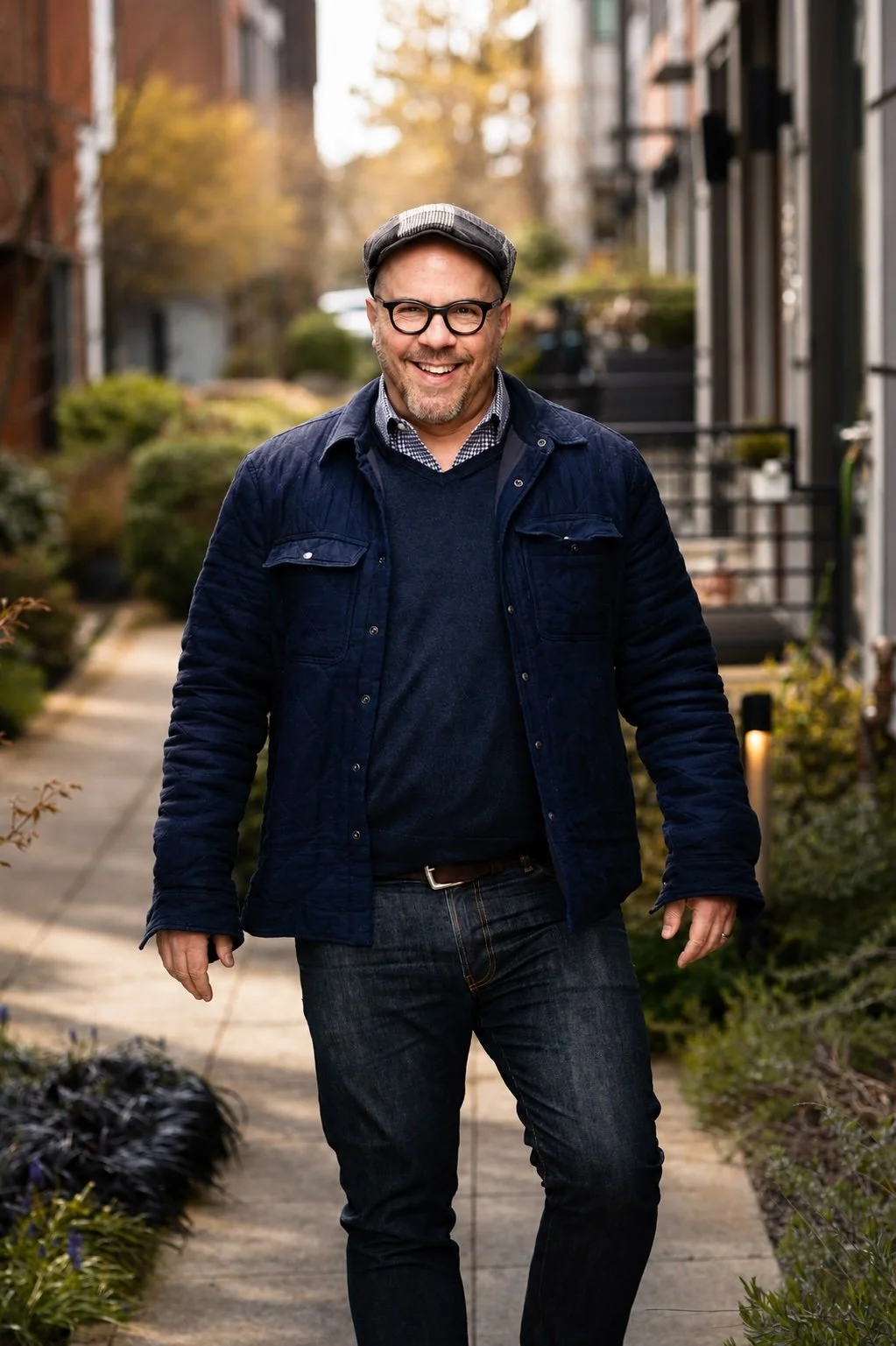 Matthew Richmond walking toward camera wearing blue jacket, sweater, jeans, check-collar shirt, with cap and glasses in an urban alley setting with landscaping.