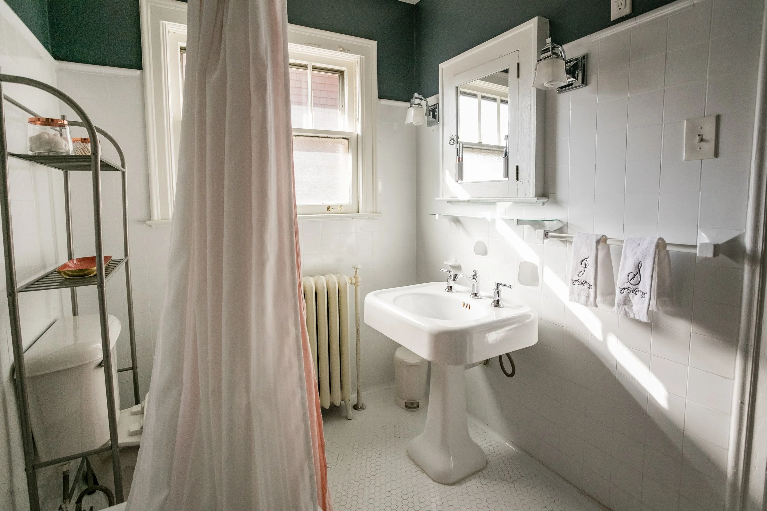A small bathroom with a white pedestal sink, mirror, and window. The window has white trim and is partly open, letting in sunlight. A white towel with embroidery hangs on a towel bar. A shower curtain partially obscures a shower or bathtub. There is a metal shelving unit with a dish and decorative items. The walls are tiled half up, with a dark upper section, and the floor has small white hexagonal tiles.