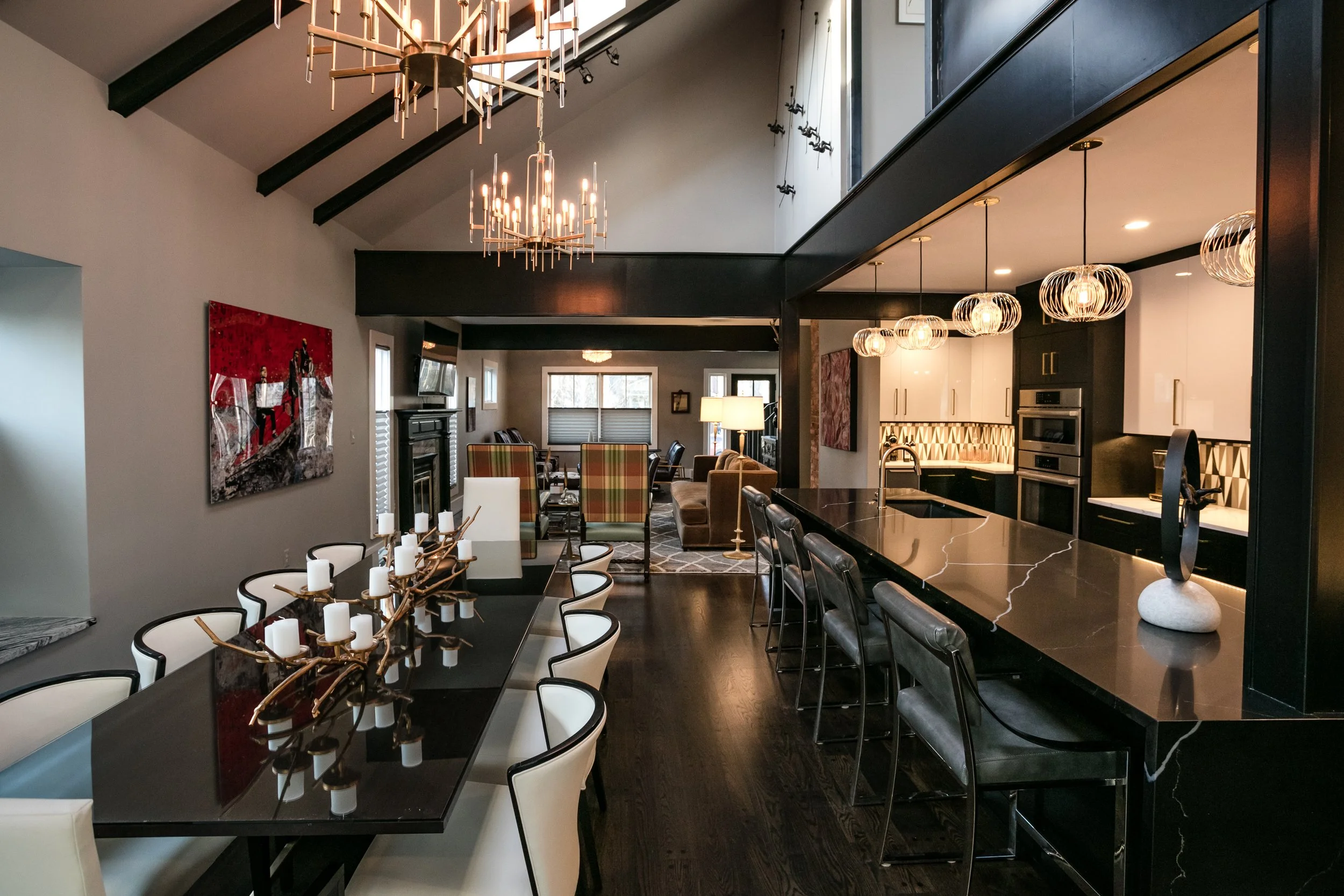 Reimagined Detroit area 1922 modern craftsman open-concept interior of a house with a dining and living area, featuring black ceiling beams, contemporary chandeliers, and a black and white kitchen with pendant lights.