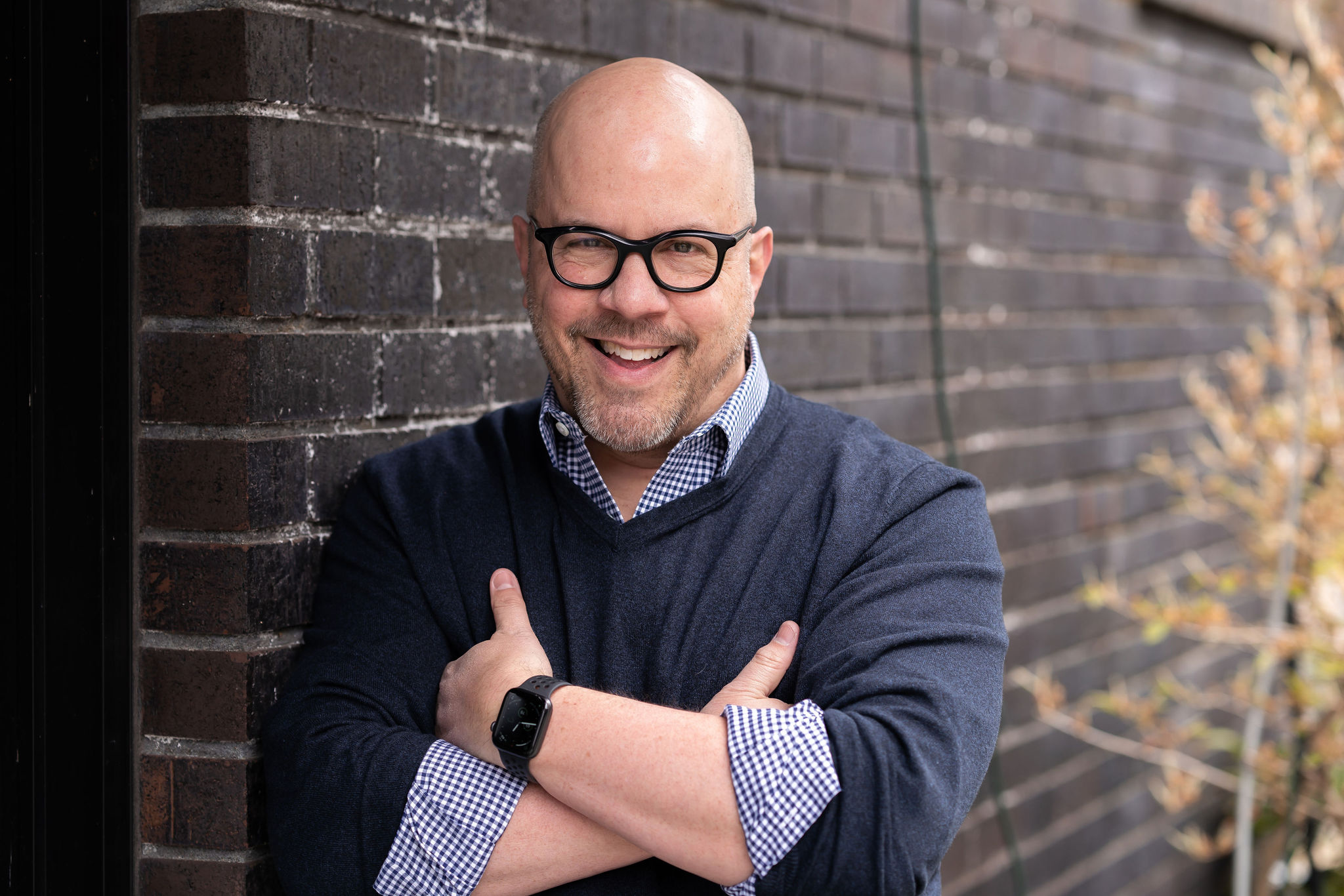 Matthew Richmond, Seattle historic home Realtor headshot wearing glasses, a checkered shirt, and a dark sweater, standing against a brick wall with arms crossed.