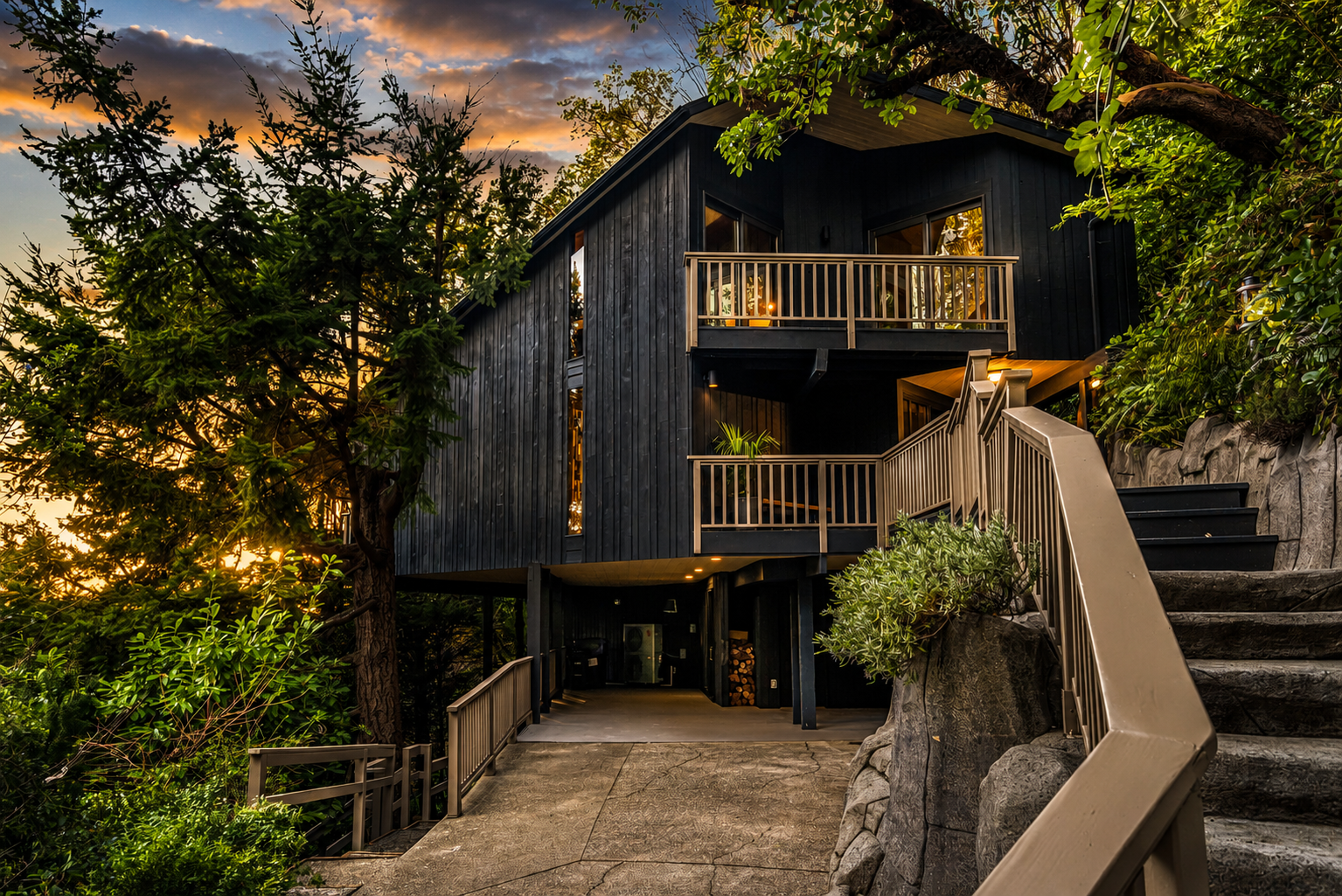 Renovated 1966 PNW contemporary in Seattle area, modern black house with multiple balconies, set among trees and rocks, during sunset with illuminated interior lighting.
