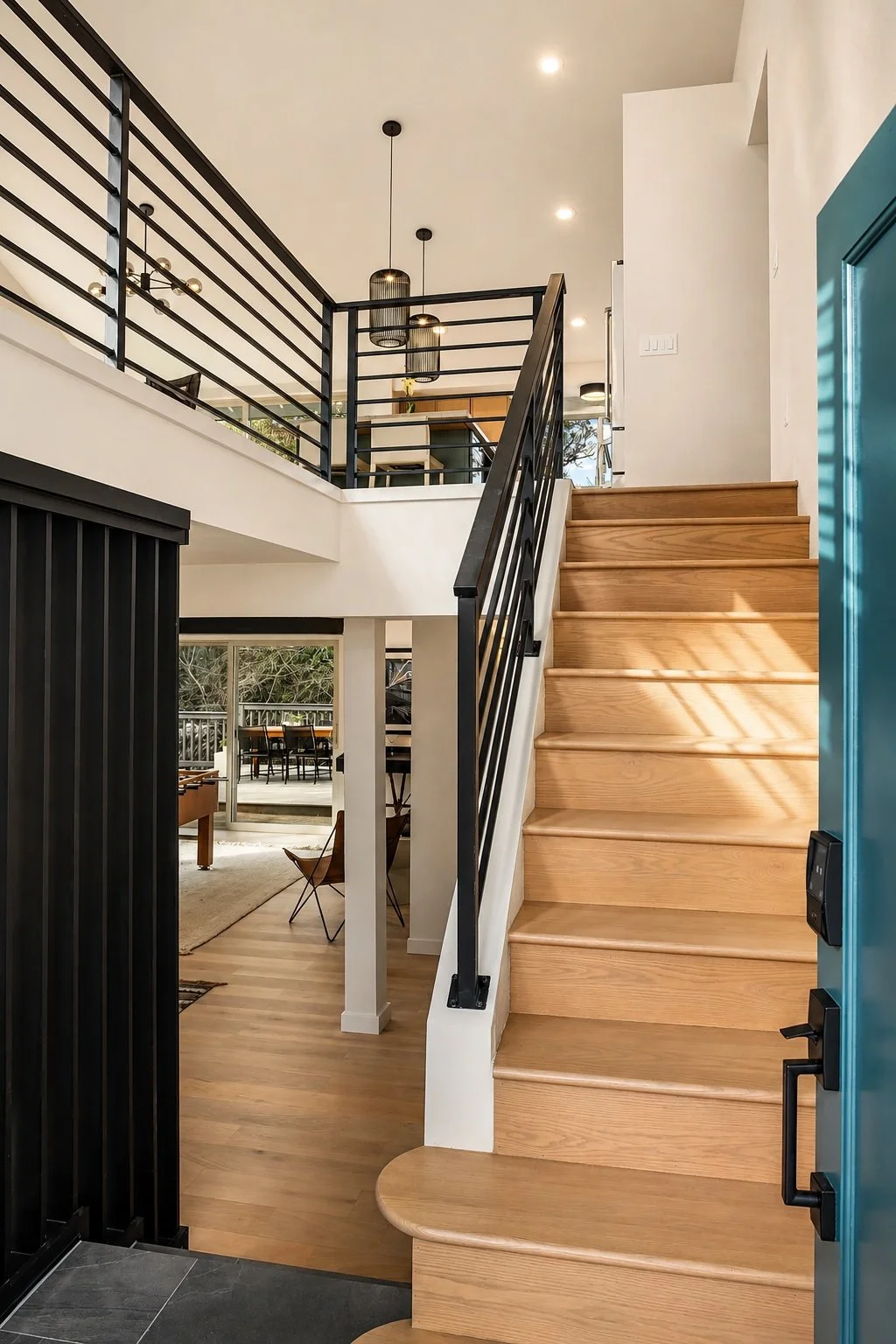 Entry of a 1970s split-level PNW contemporary home with light oak wood stairs and floors, a black metal railing with horizontal rails, a vaulted ceiling above, black slats on left, and turquoise open entry door open with black handle on the right.