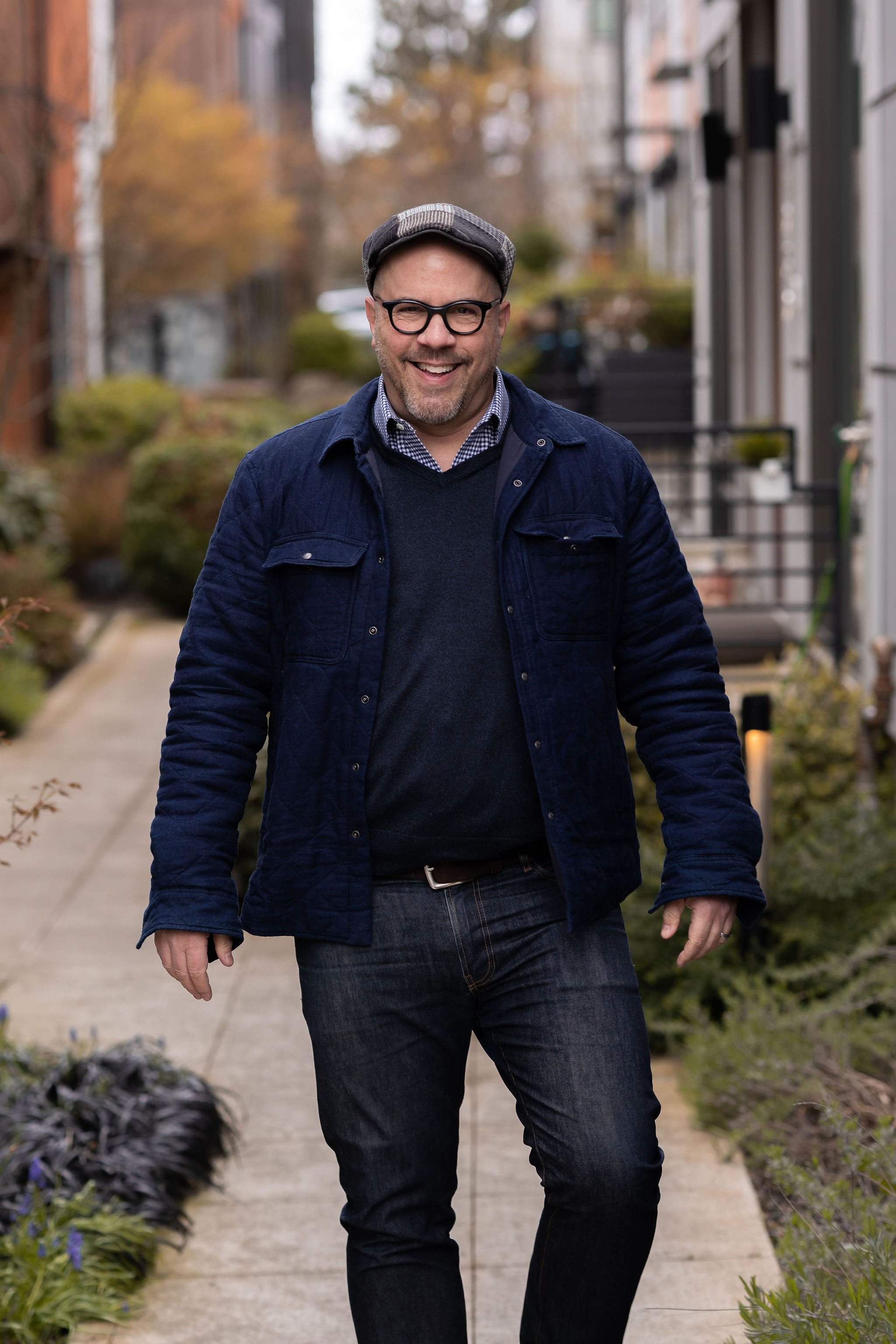 Matthew Richmond, who represents buyers and sellers of historic Seattle property, smiling and walking outdoors, wearing glasses, a flat cap, a blue jacket, and jeans, with blurred autumn trees and residential buildings in the background.