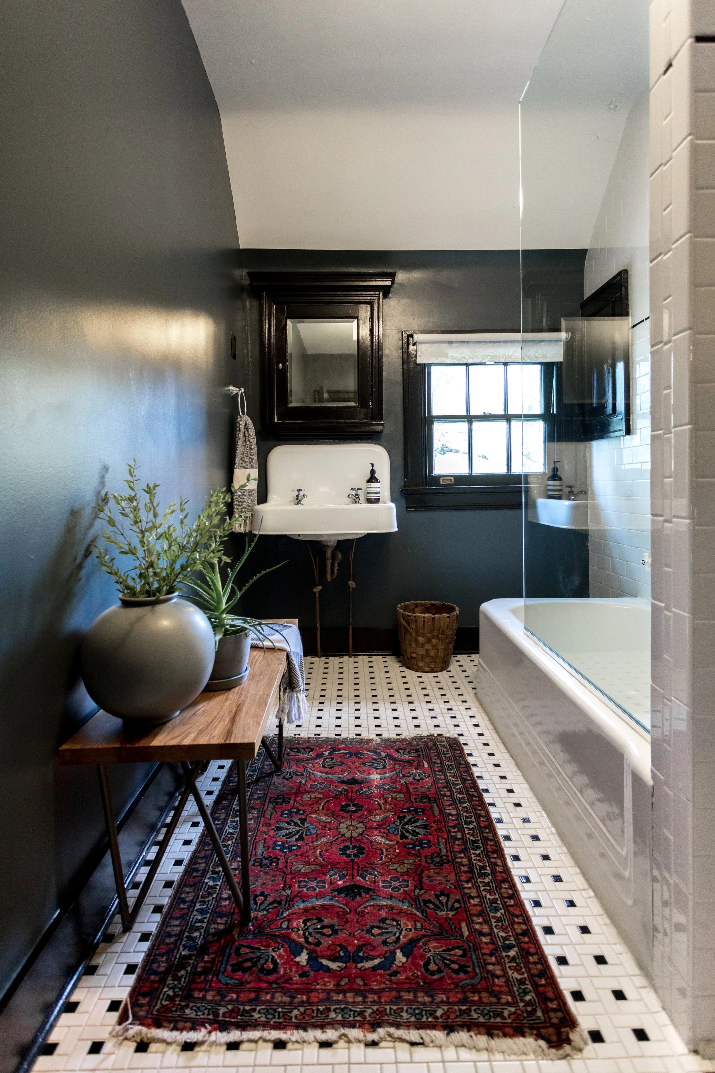A vintage-style bathroom with a dark wall, black framed mirror above a white sink, window with white shade, glass shower door, white subway-tile shower wall, a wooden table with green plants in beige vases, woven basket, red patterned rug, and black and white tile floor.