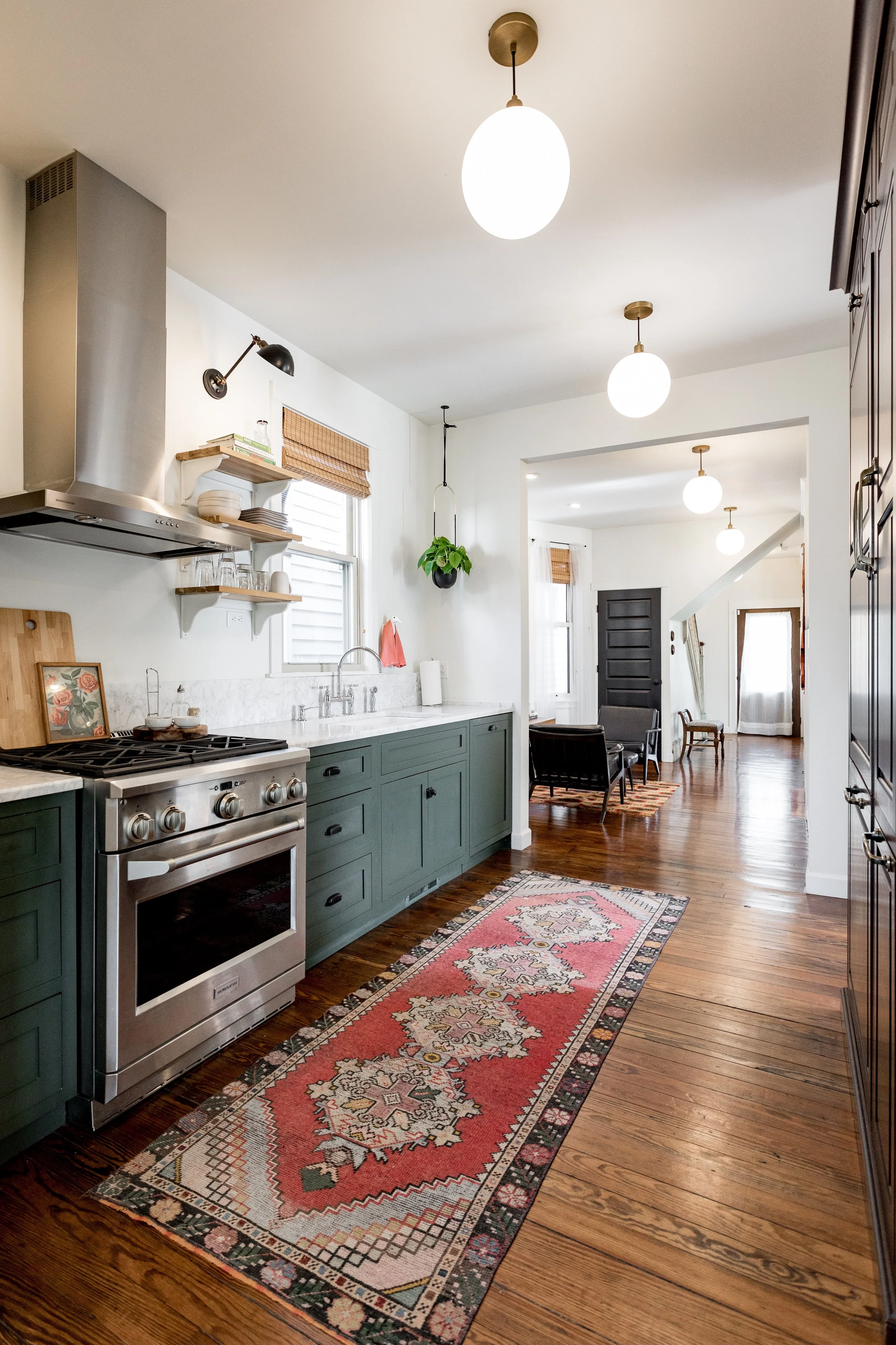 Reimagined 1897 Detroit Victorian kitchen with green cabinets, stainless steel stove, open shelving with dishes, a window with blinds, hanging plant, and a living room area with chairs, a rug, and a dark front door in the background.
