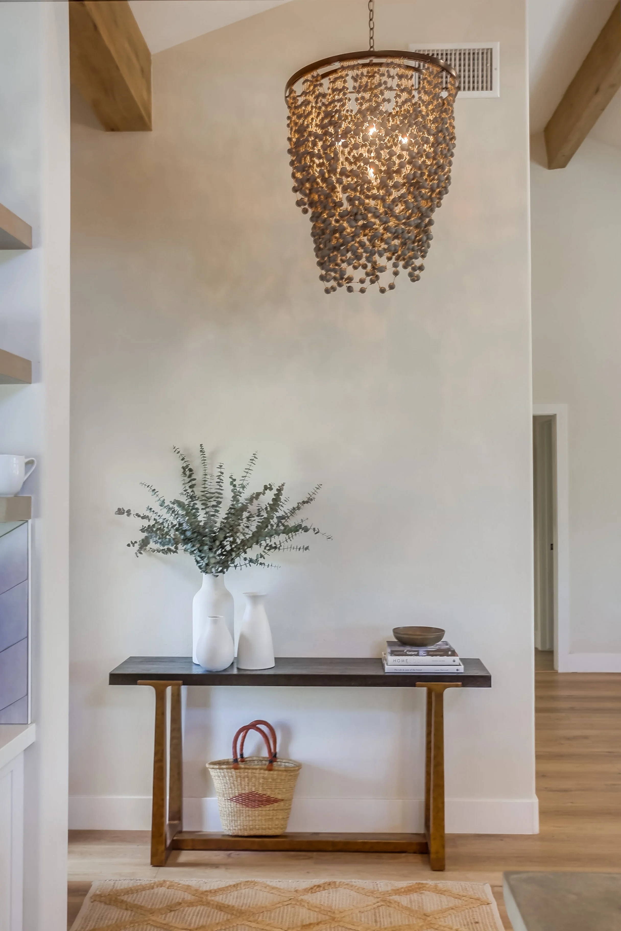 entryway styling with wood console, decorative accents, and layered decor in San Diego home.  light oak hardwood floor. A decorative chandelier hangs from the ceiling, and beige walls with wooden beams are visible.