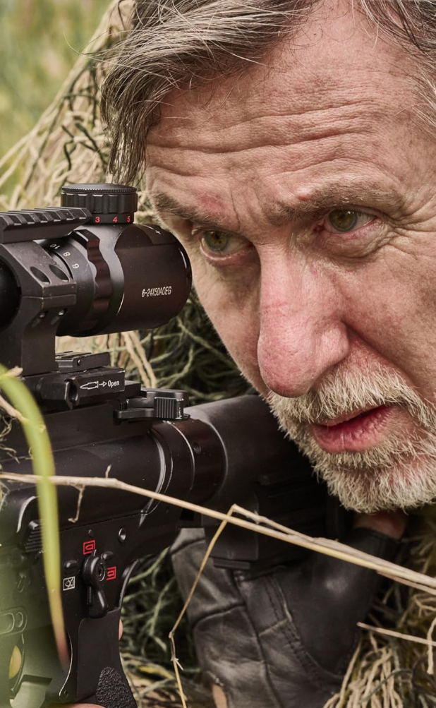 Tim Roth (Academy Award Winner) in Seven Snipers Feature Film Releasing in Cinemas in April - Upcoming Action Movies In Cinemas - Close-up of a man aiming through a scope with one eye, lying low in tall grass.