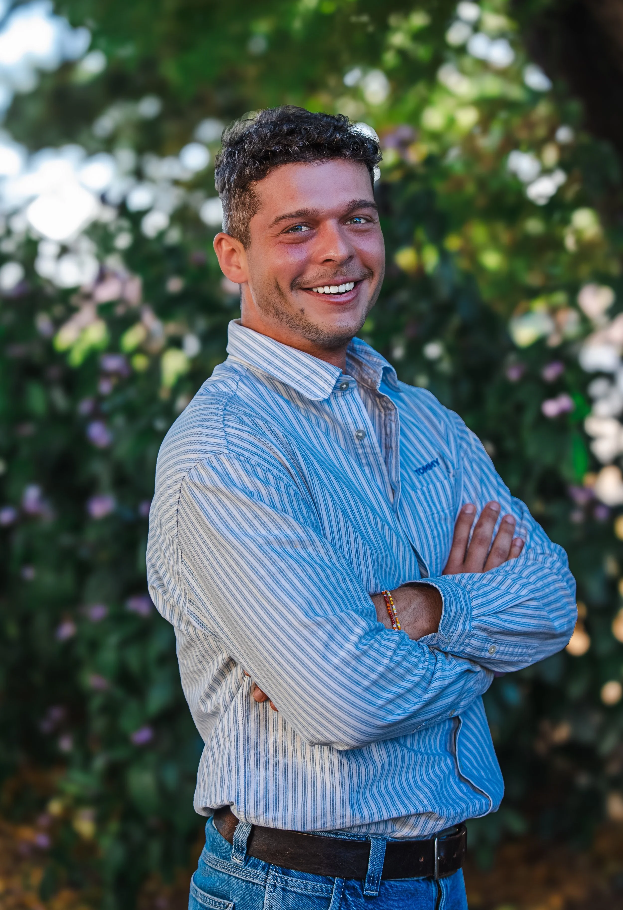 Hugo Bouët, social media manager, souriant avec des bras croisés, portant une chemise à rayures bleues, devant un fond flou de nature avec des plantes et des fleurs.