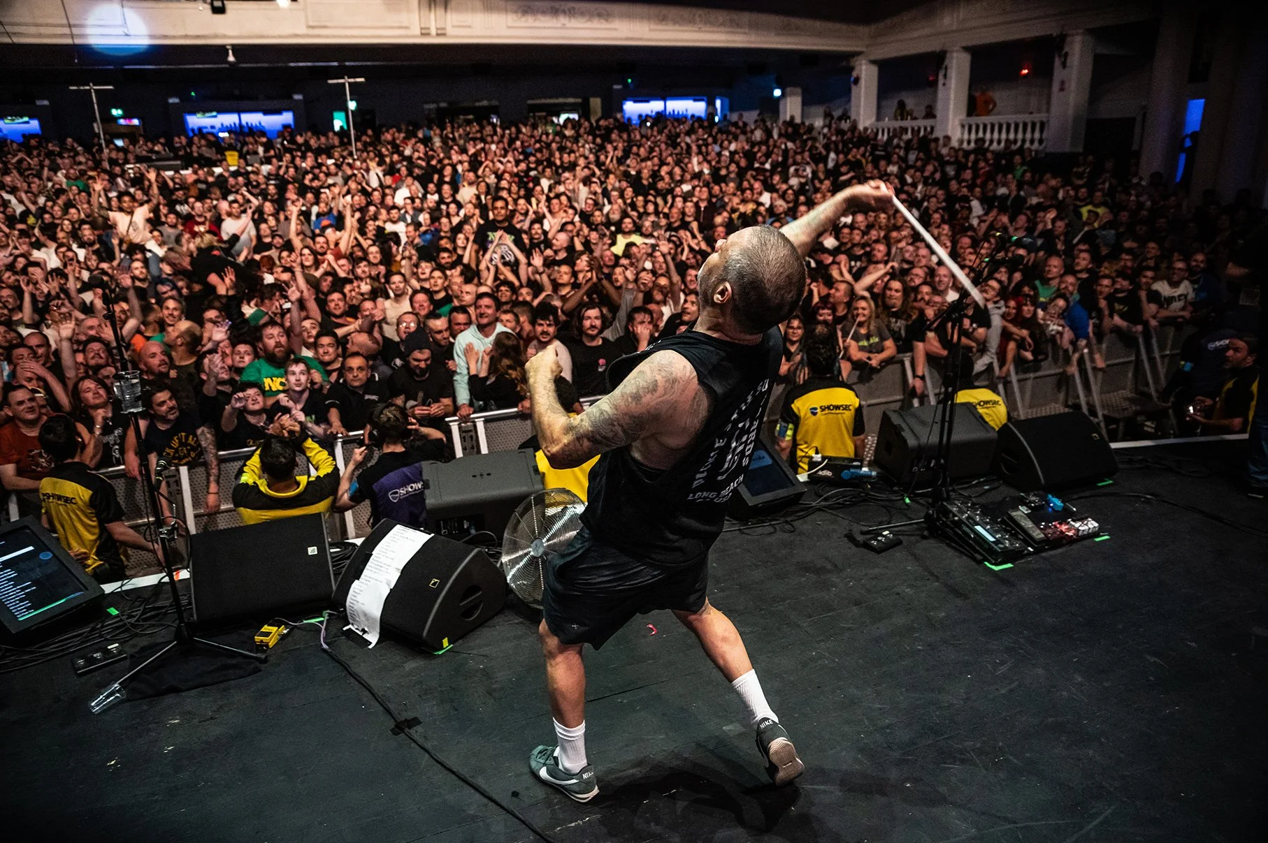 A performer with tattoos on his arms, wearing a black sleeveless shirt, black shorts, white socks, and sneakers, is energetically dancing on stage in front of a large crowd at a concert or live event.