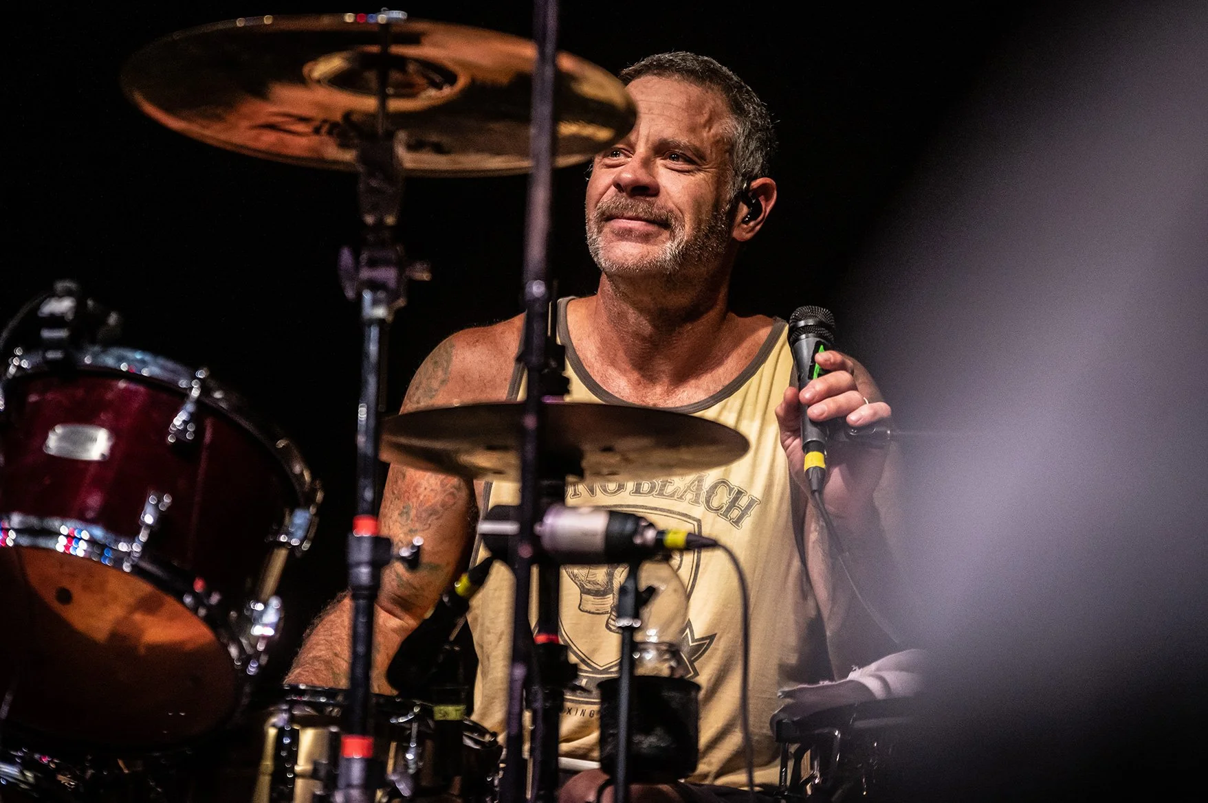 A man with short gray hair and a beard, wearing a beige tank top, appears to be singing into a microphone while sitting behind a drum set during a performance.