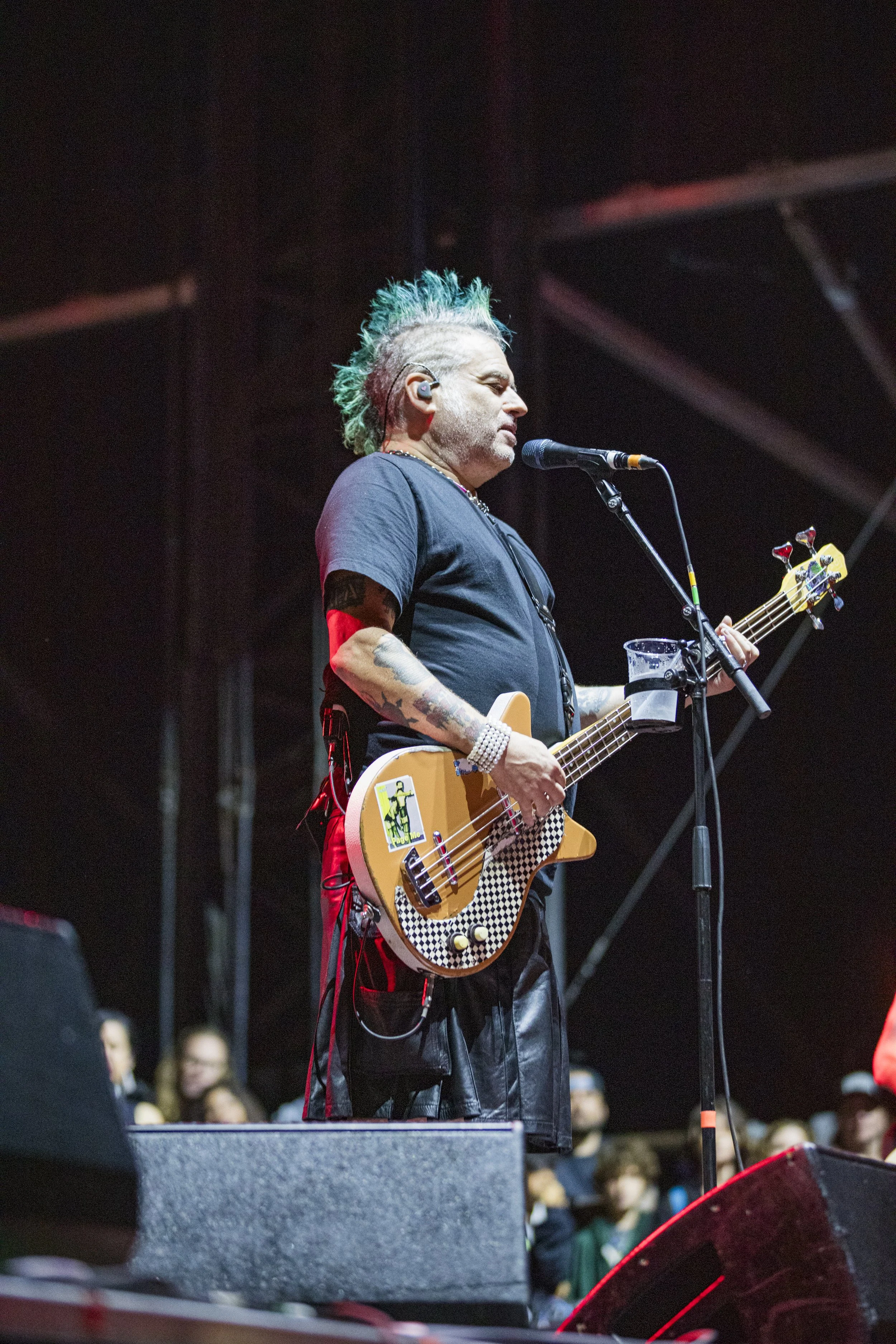 A musician with a mohawk hairstyle, tattoos, and wearing a black t-shirt, playing a bass guitar on stage during a concert. There are people in the background watching the performance.
