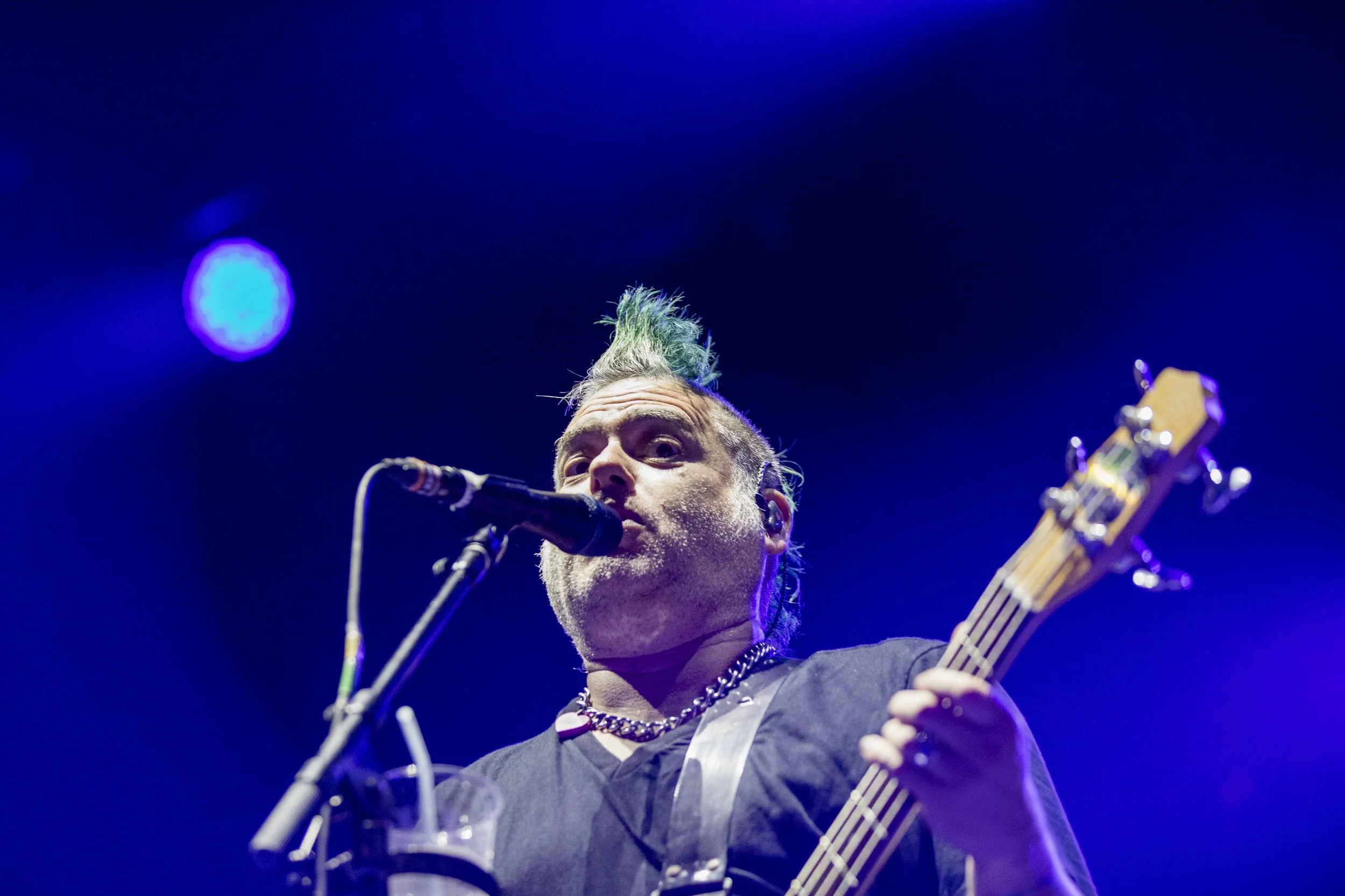 Male musician with green mohawk hairstyle playing guitar and singing into a microphone on stage with blue lighting.