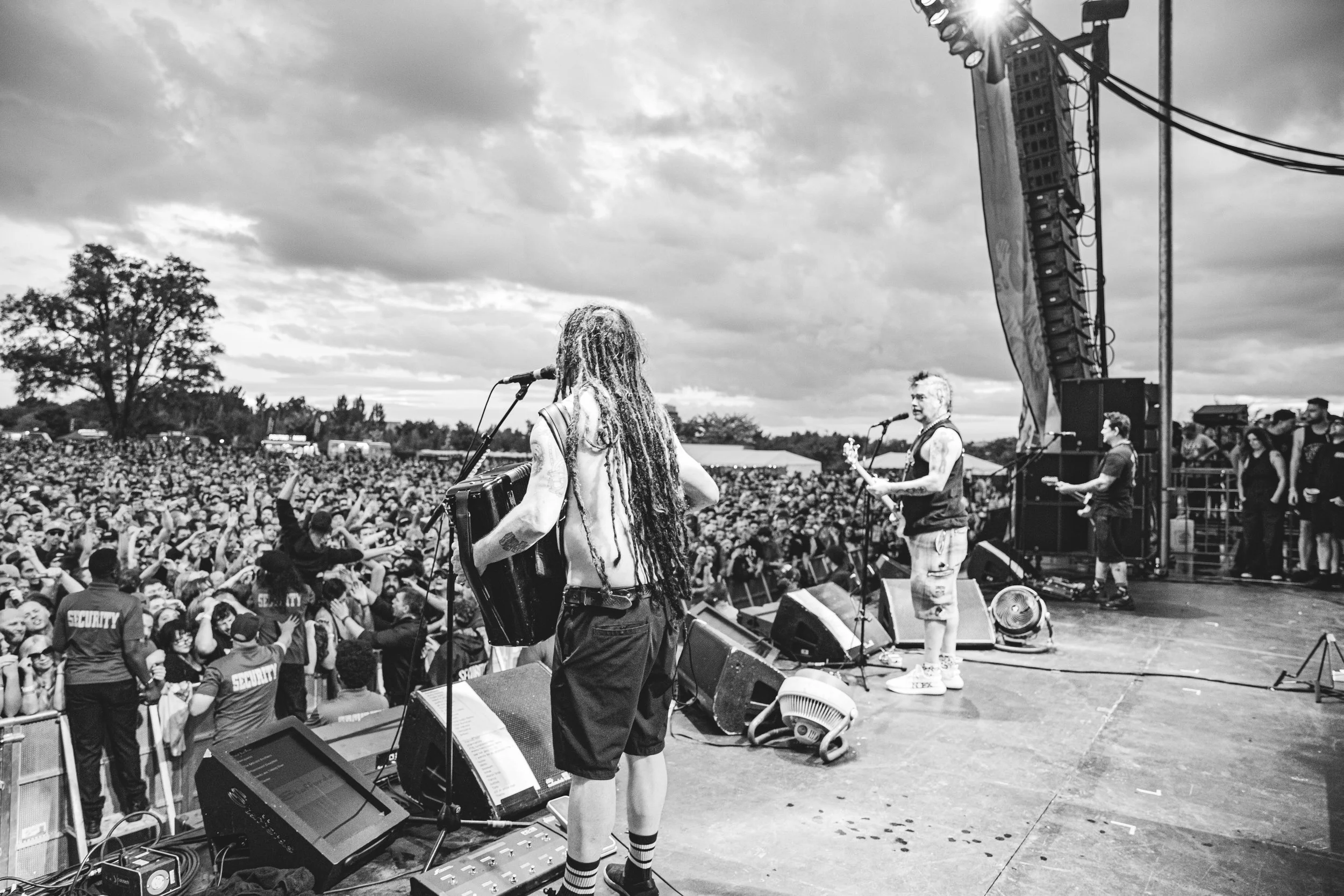 Black and white photo of a band performing on stage at an outdoor concert with a large crowd watching under cloudy skies.