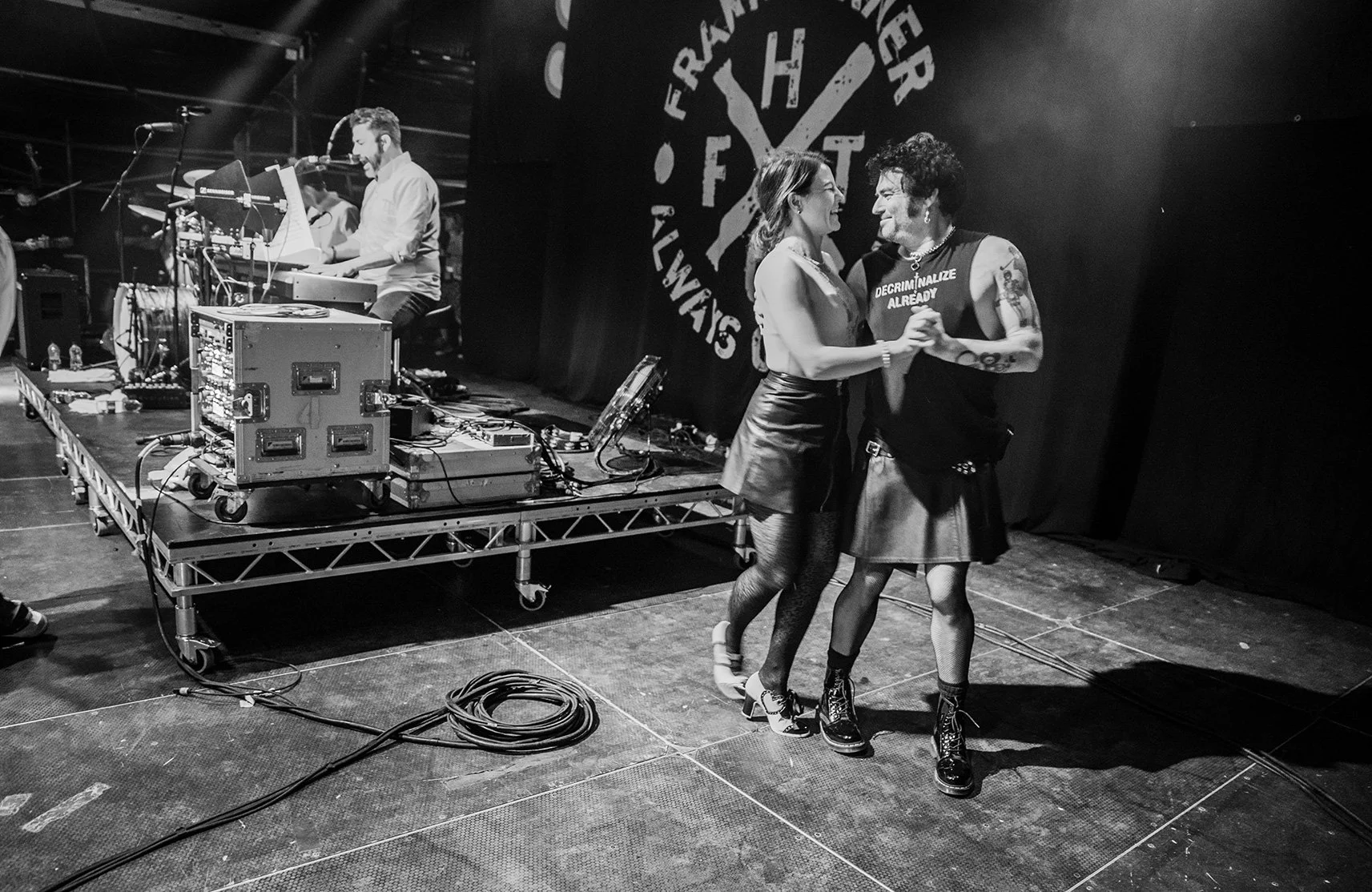 Two women dancing close together in front of a band playing on stage. The women are smiling, holding hands, and wearing dark clothing. The band members are visible in the background, with musical equipment and a large logo on the back of the stage.