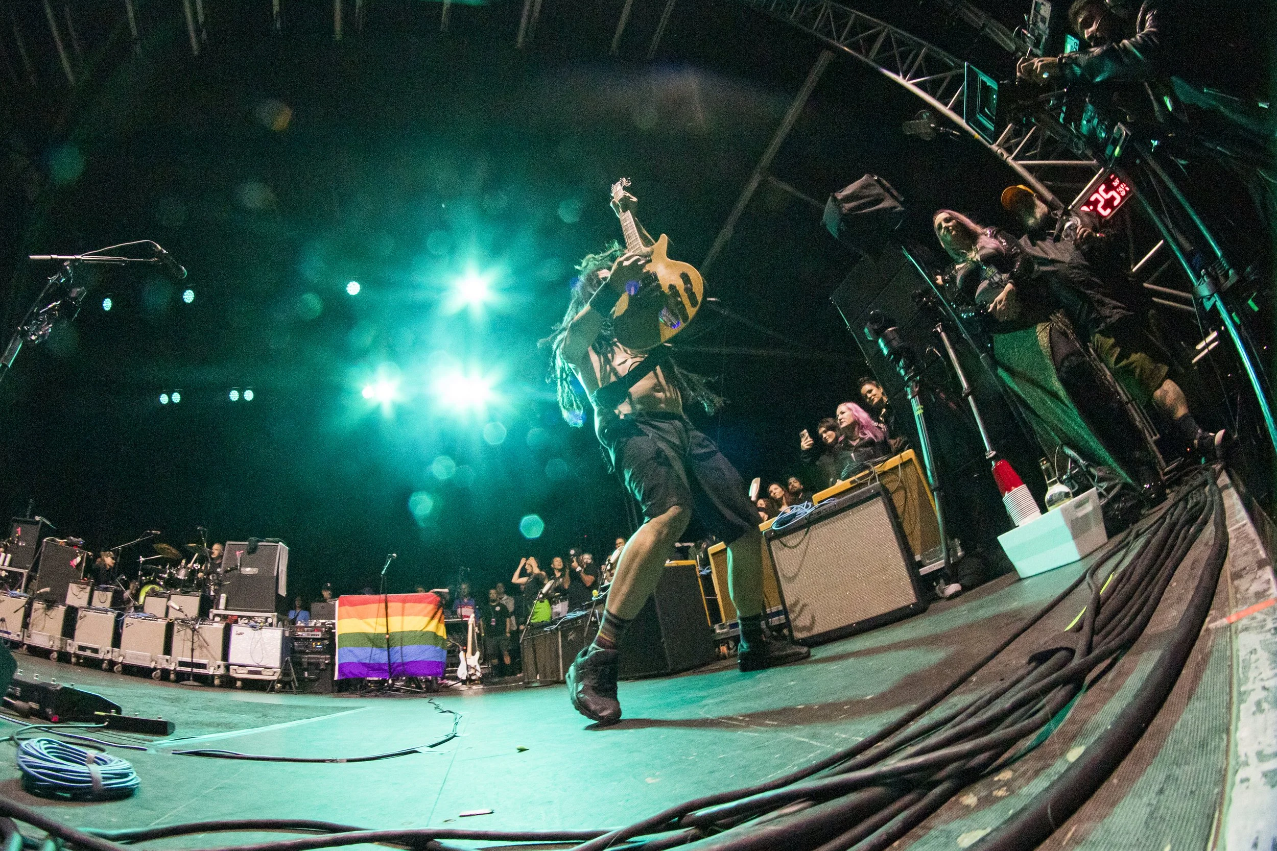 A performer with dreadlocks on stage holding an acoustic guitar above their head during a concert. Bright stage lights shine from behind, with a crowd and on-stage equipment visible in the background.