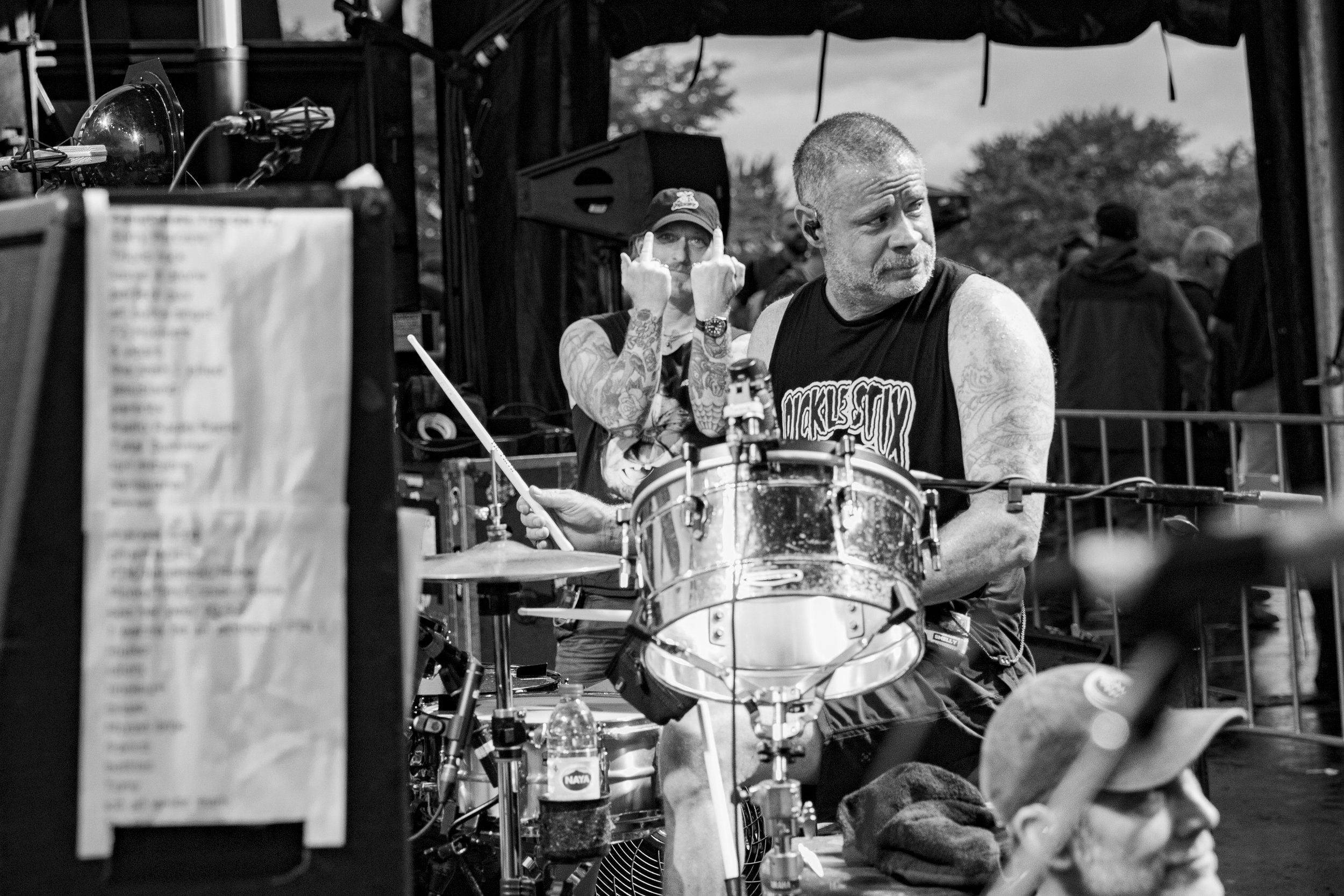 Two men with tattoos, one playing drums and the other in the background making a rude gesture with middle fingers, during an outdoor event.
