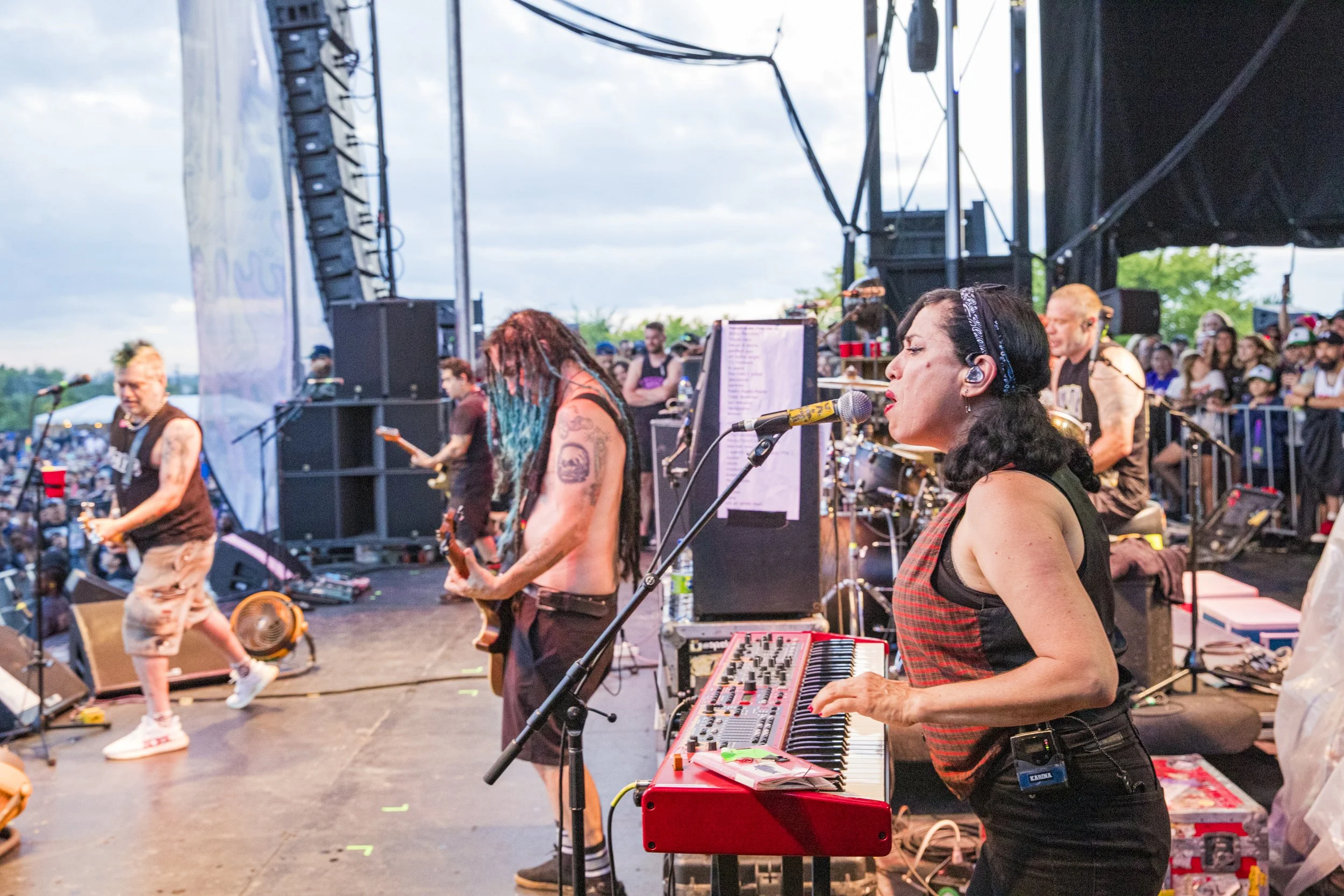 A live outdoor concert with a band performing on stage, including a female keyboardist singing into a microphone, a guitarist, and other band members, with an audience in the background.