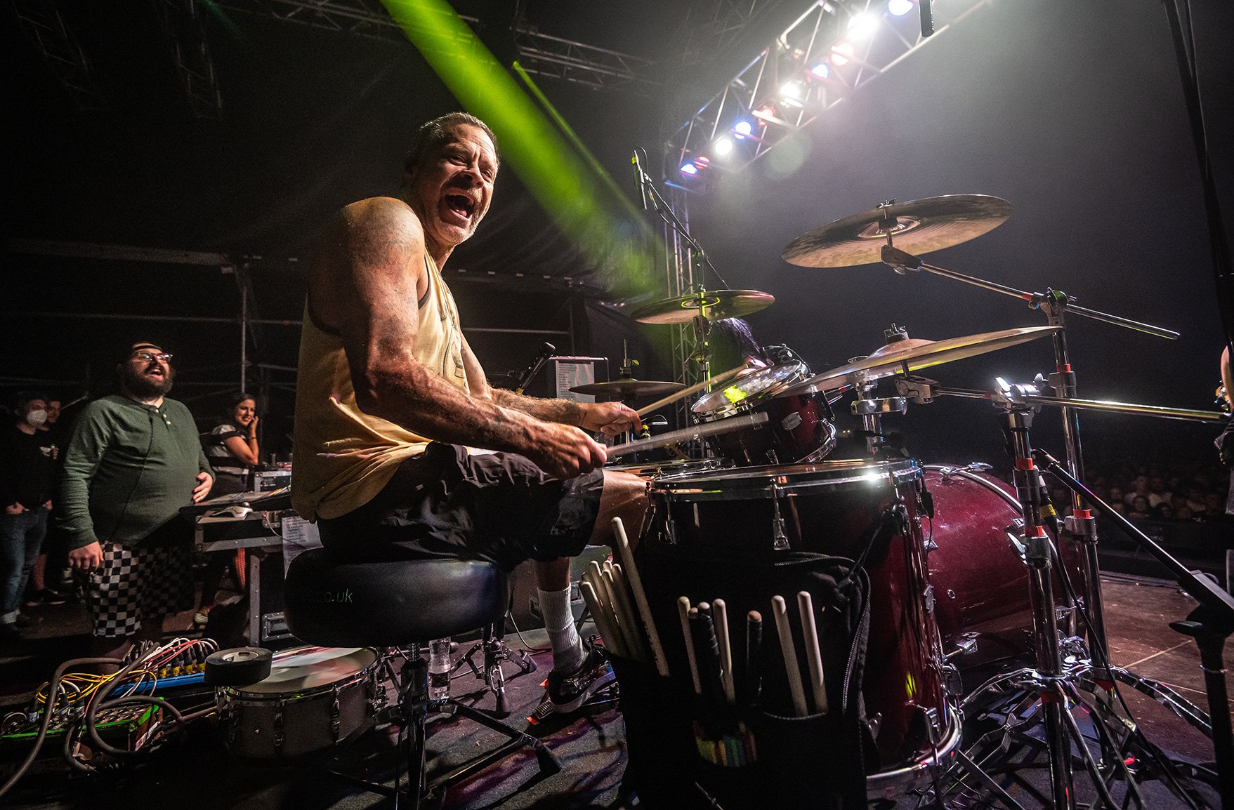 A musician passionately playing drums on stage with colorful stage lights and an audience in the background.