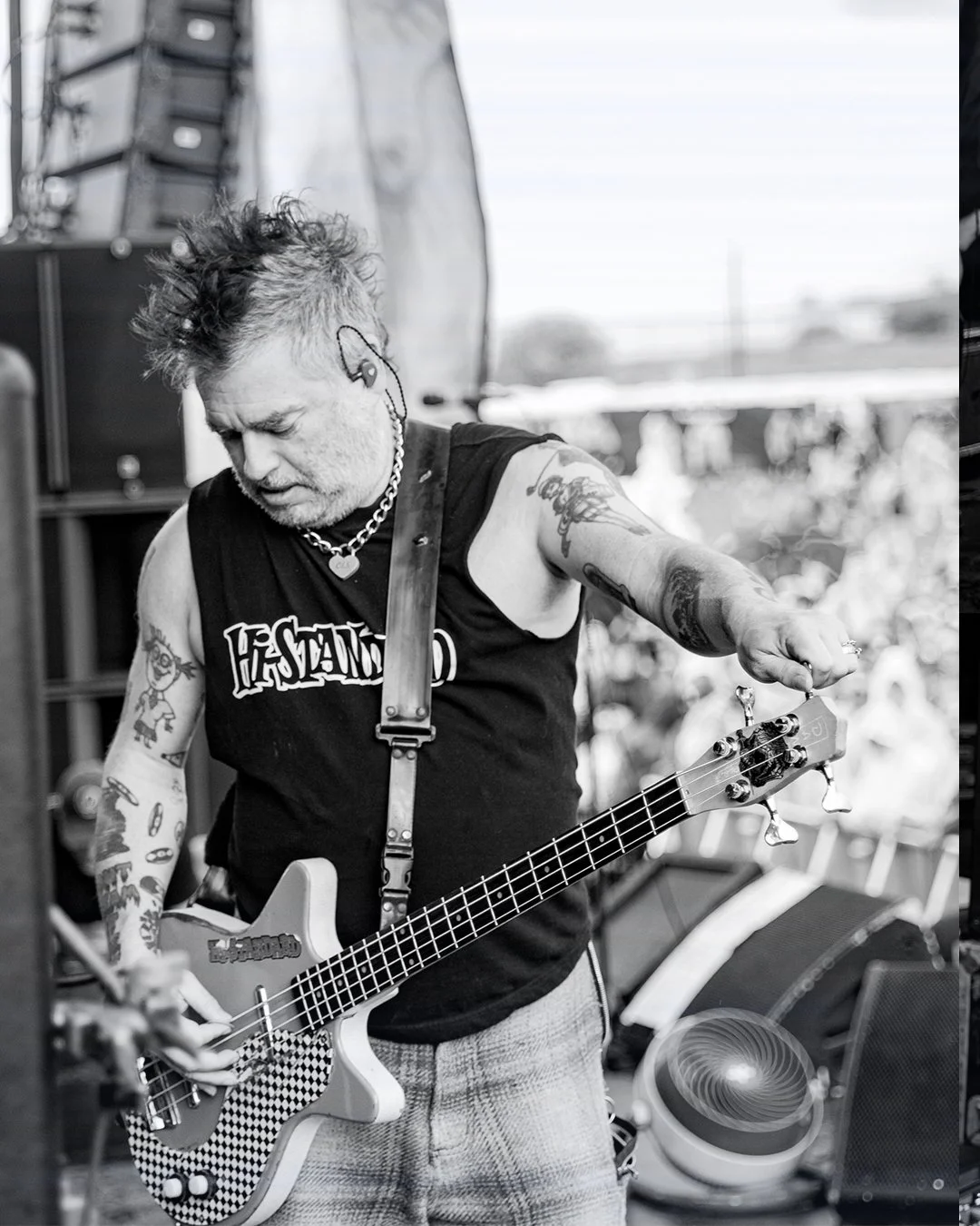 A musician with tattoos, spiked hair, and earphones in one ear, playing a bass guitar during a performance. He is wearing a sleeveless shirt, a chain necklace, and appears focused on his instrument.