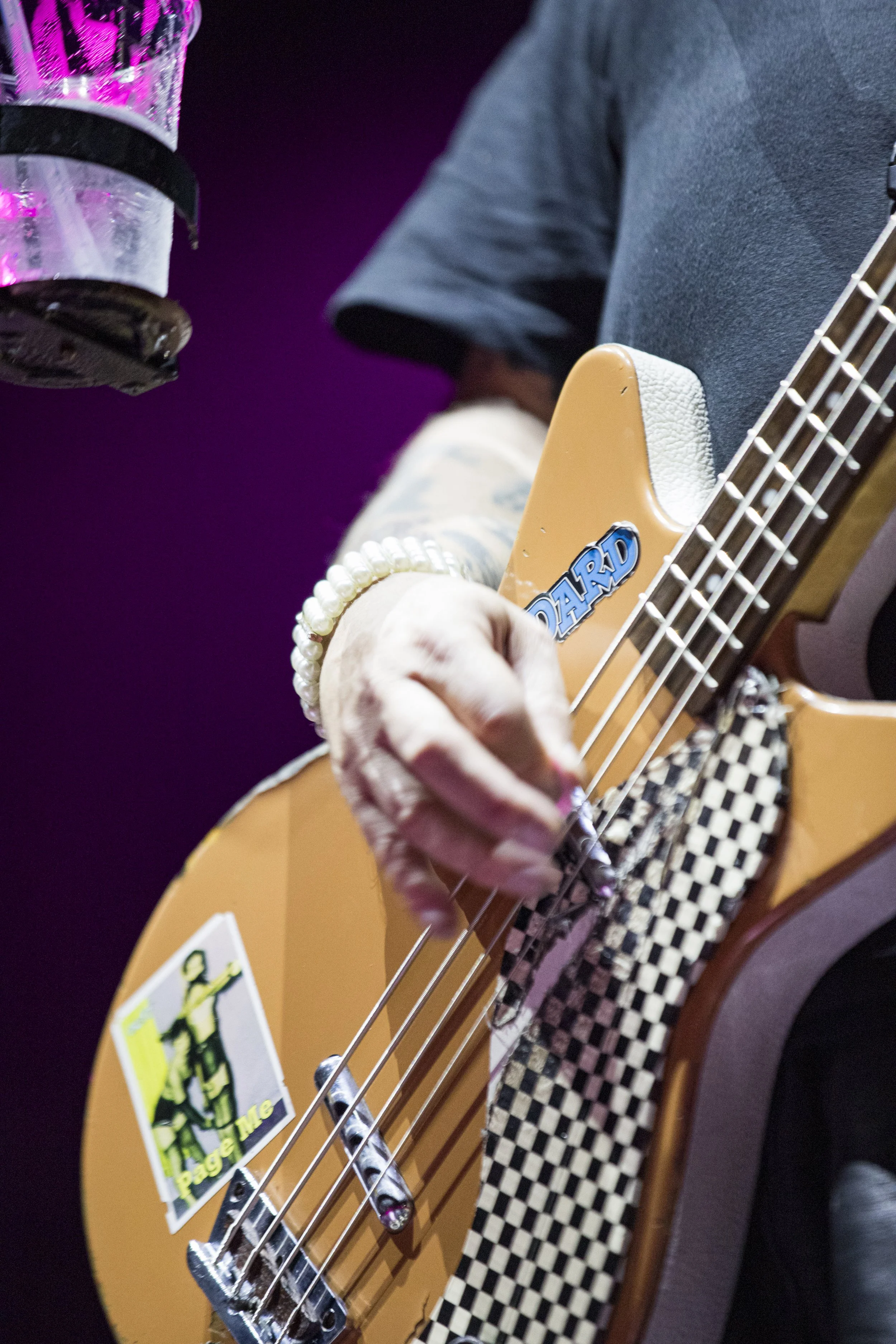 A person playing a yellow bass guitar with a sticker that says 'Tag Me' and another sticker with the word 'BOARD.' The person is wearing a dark T-shirt, a pearl bracelet, and has checkered fabric near the guitar. The background is dark and purple.