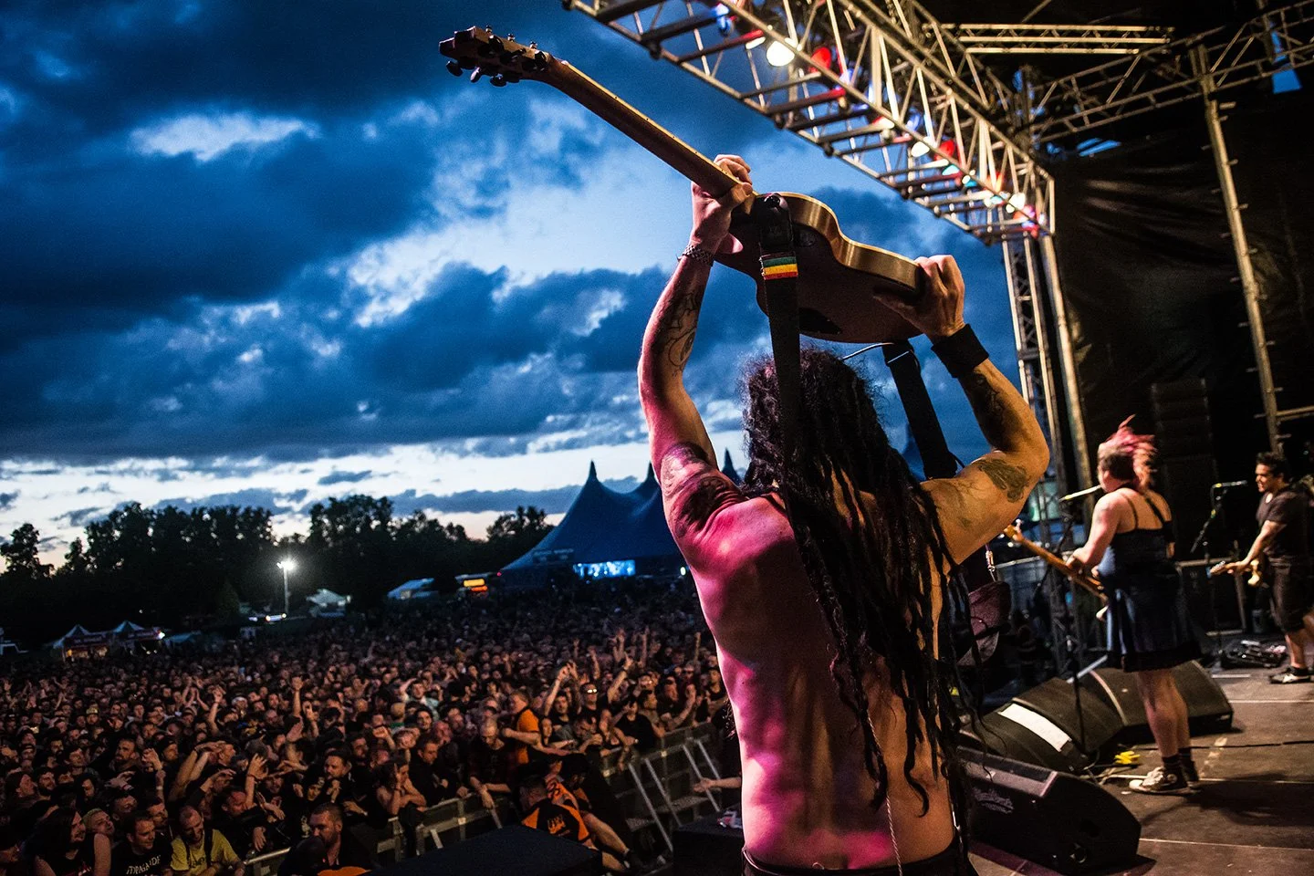 Performing musician with long dreadlocks holding a guitar above their head on stage during a concert at dusk, with a large crowd in front and a cloudy sky in the background.