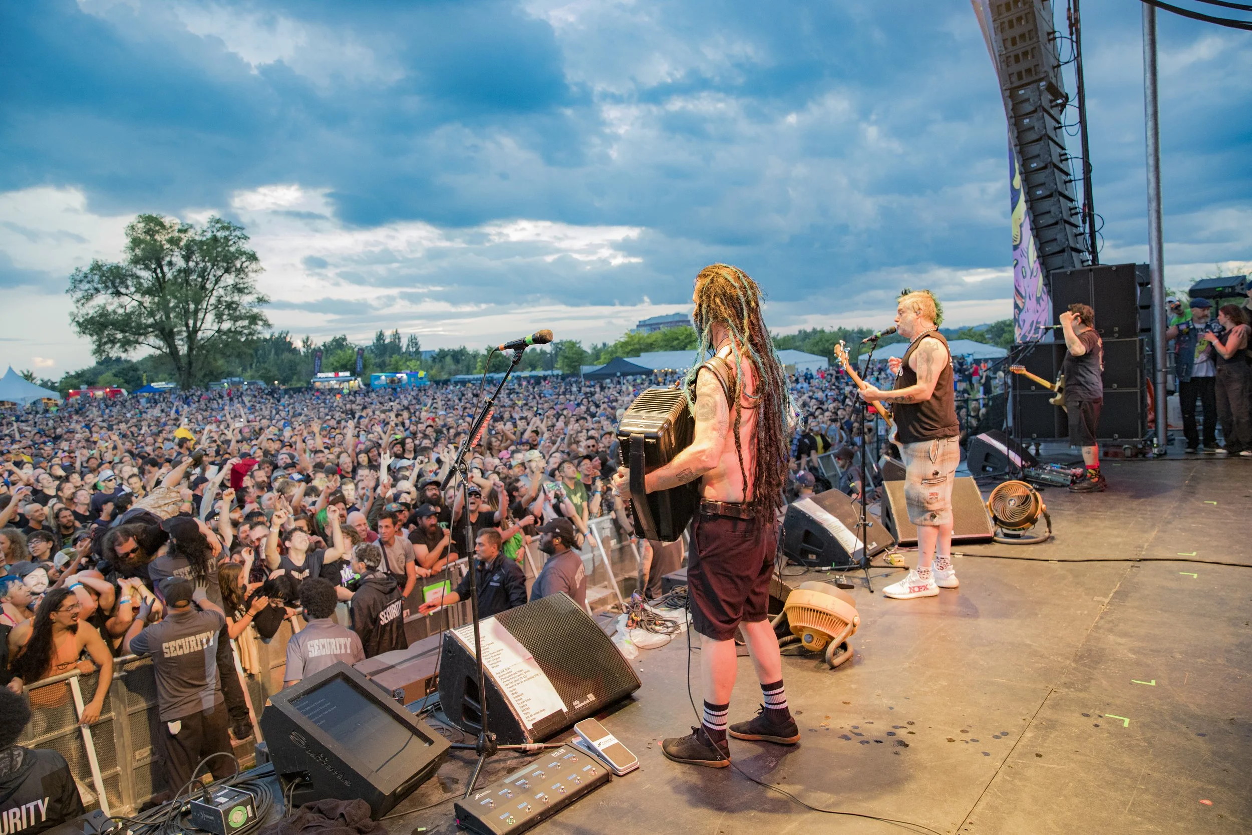 Musicians performing on stage at an outdoor concert with a large crowd under a cloudy sky.