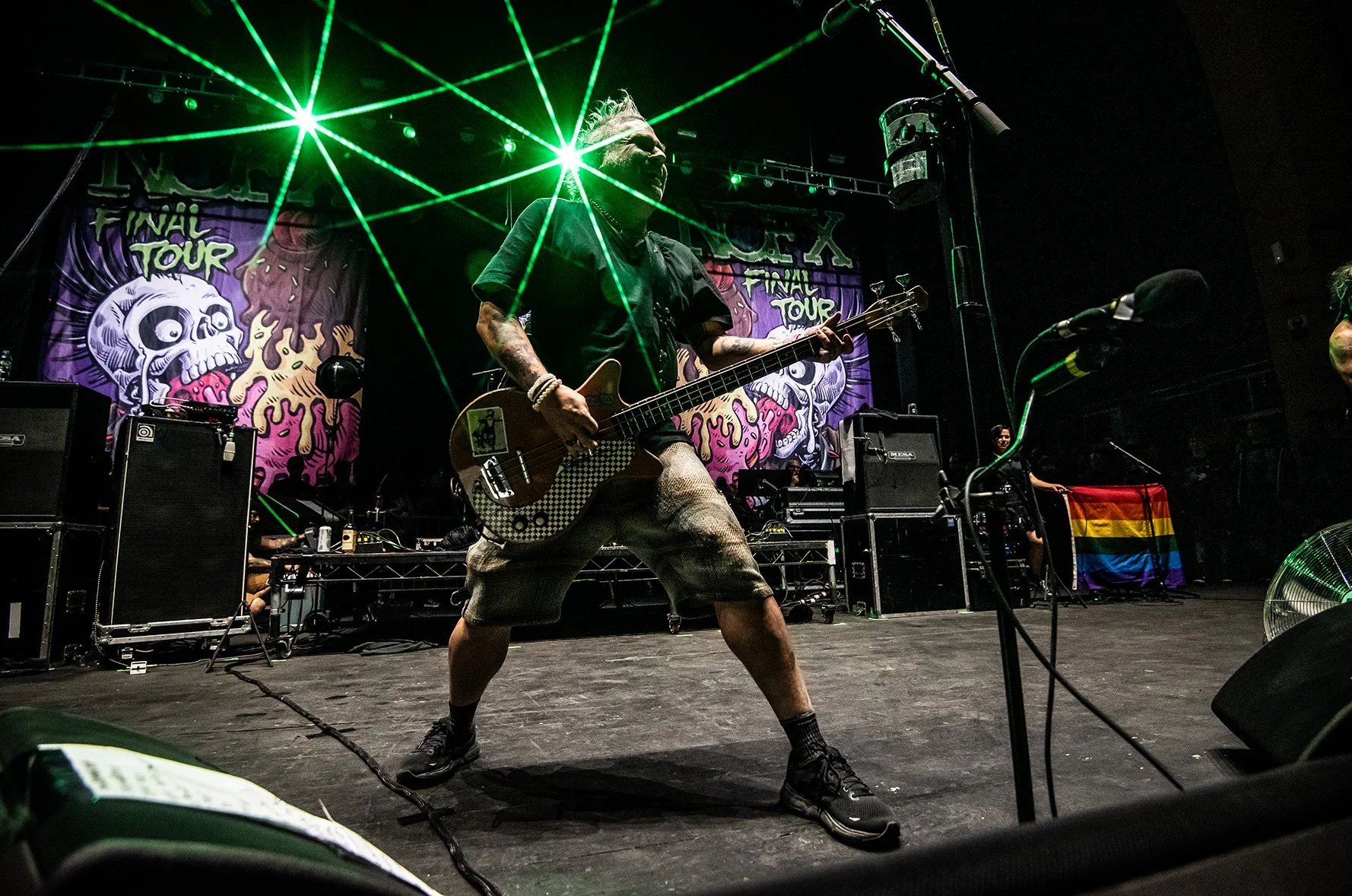 A musician playing an electric bass guitar on stage during a concert, with green laser lights and a colorful backdrop featuring cartoon skulls and text that reads 'FINHLU TOUR'.