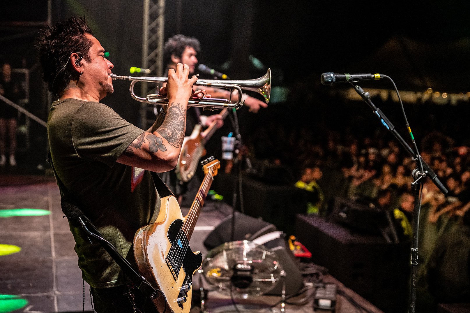 Musician playing trumpet on stage, with band members and audience in the background during a concert at night.