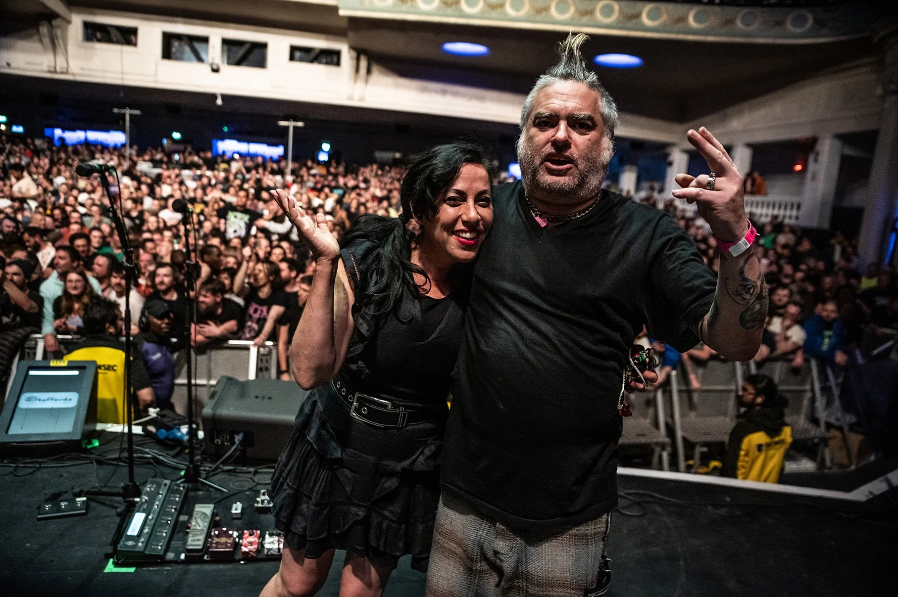 Two performers on stage in front of a large, seated audience; both are smiling and making hand gestures, with the crowd and stage equipment visible in the background.