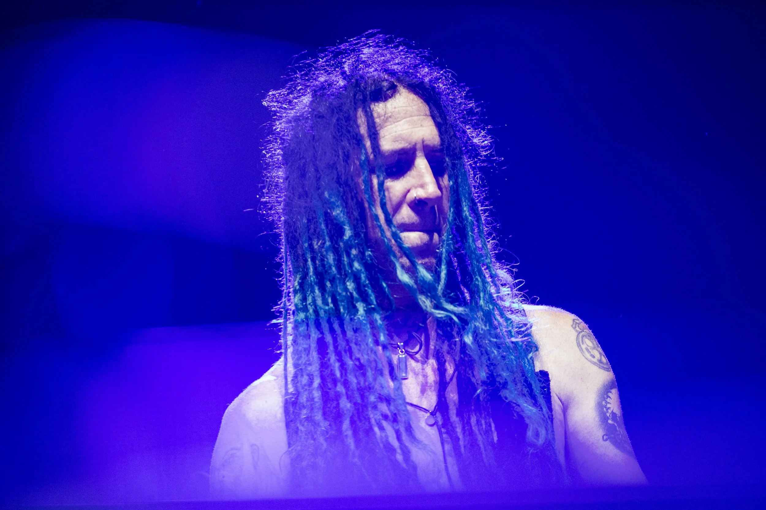 A person with long dreadlocks, some dyed blue, on stage under purple lighting, looking down with a serious expression, wearing a necklace and tattoos visible on arms.